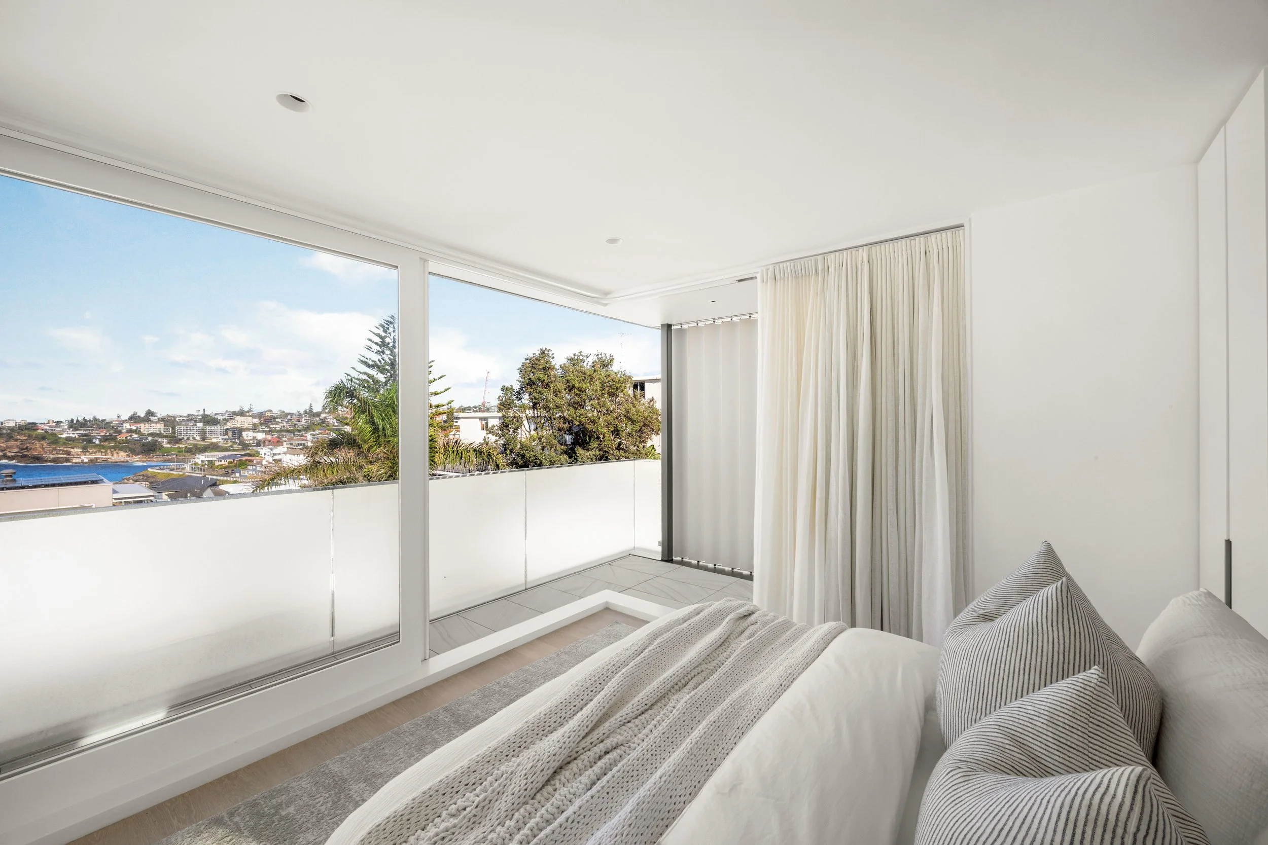 Modern bedroom with large glass sliding door, white curtains, and a view of a cityscape with trees and buildings.