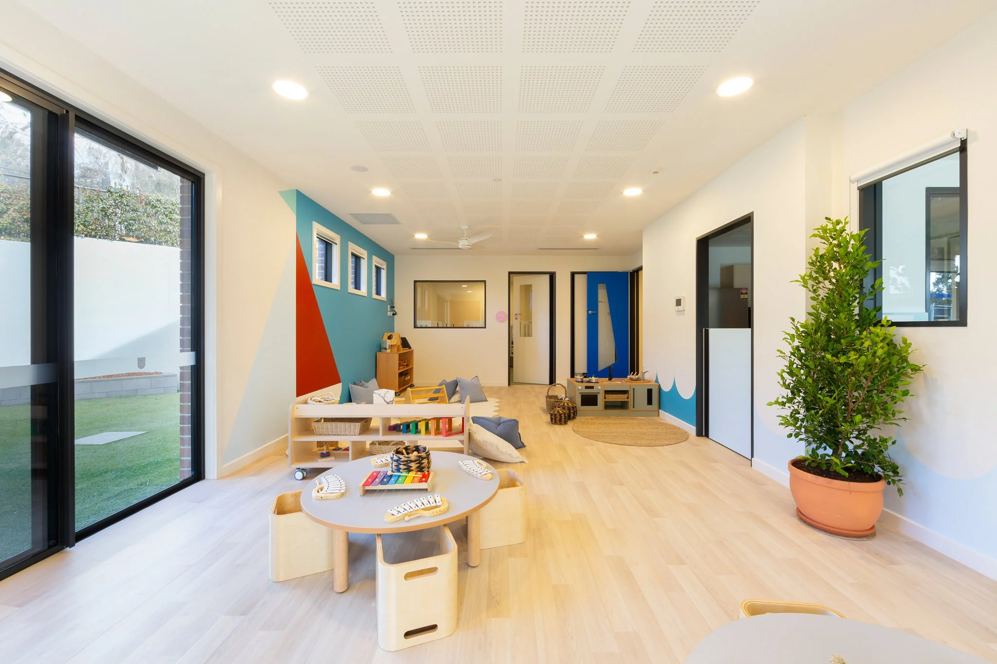 Brightly lit playroom with large sliding glass doors, a colorful geometric wall design, small table with musical instruments, and various toys and cushions on the wooden floor. Potted plant is on the right.