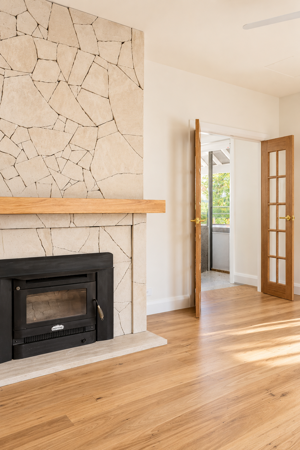 Living room with a beige stone fireplace and a wooden mantel, hardwood floors, and double wooden doors leading to a balcony with outdoor view.