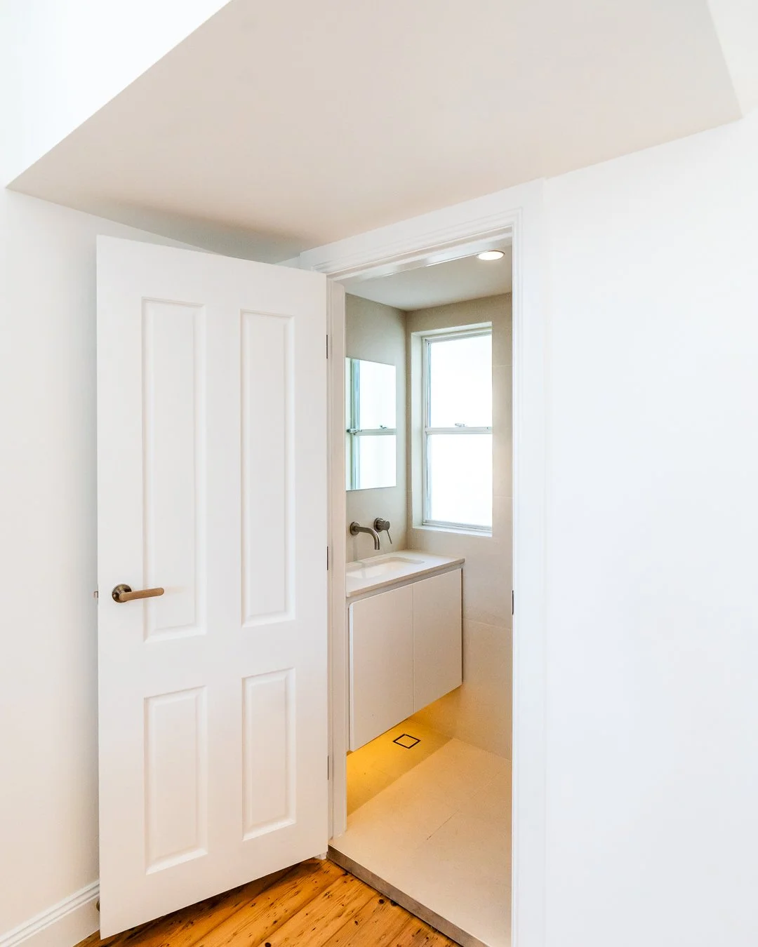 A small laundry or utility room with a white door partially open, revealing a small sink with cabinet underneath, a window, and a mirror on the wall.