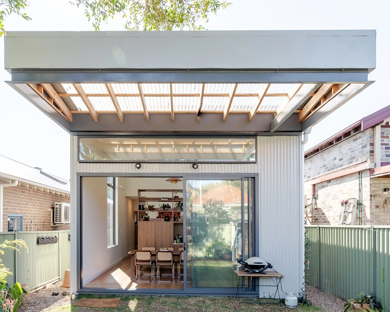 Modern small house with corrugated metal exterior, large glass sliding door, and an overhanging roof with wooden support beams. The interior features a dining table and shelves with decorative items. A small table outside holds a grill.