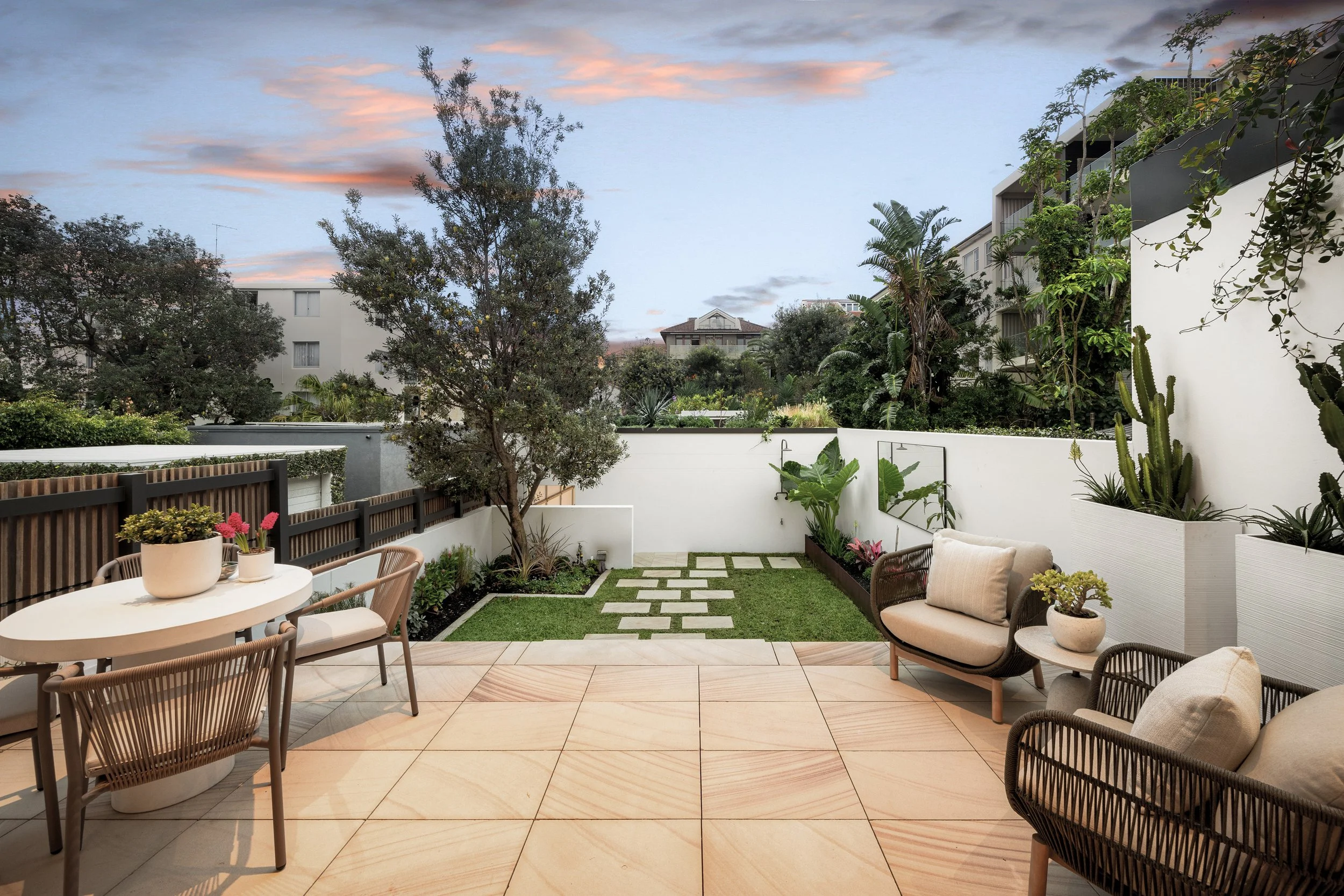 Outdoors patio with round table and chairs, cushioned seating, potted plants, greenery, trees, neighboring buildings, and a sky with scattered clouds at sunset.