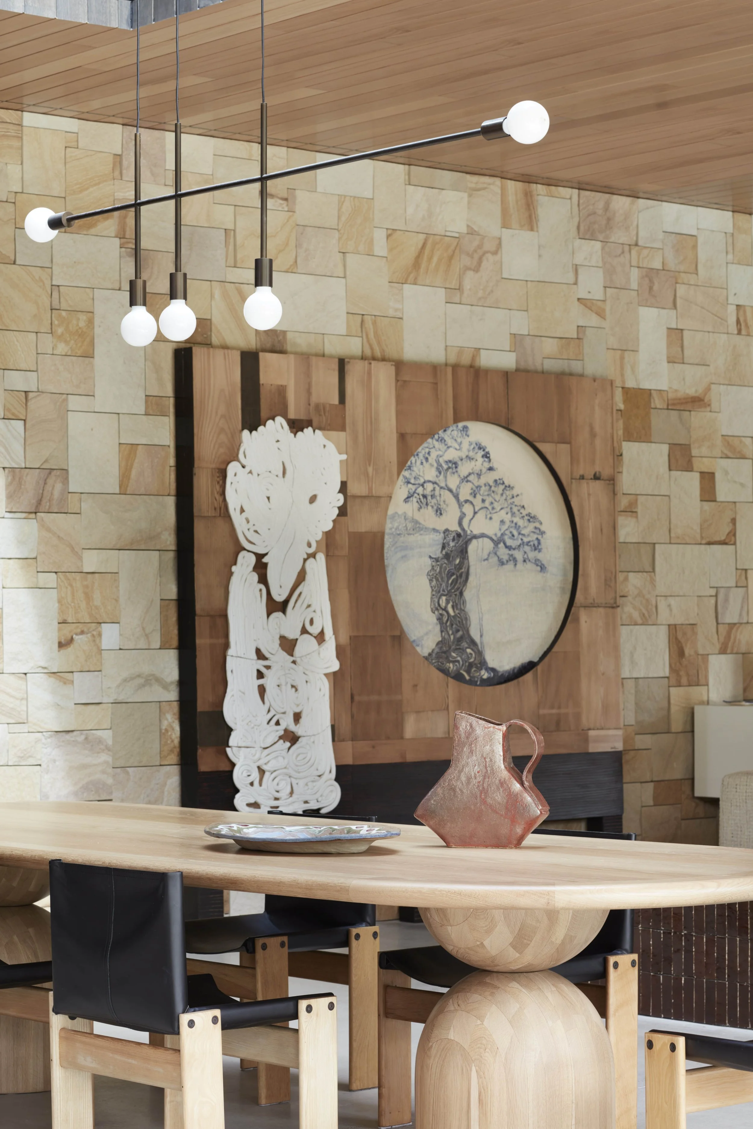 Interior of a modern room with decorative wall art, a wooden table with a ceramic pitcher, black chairs, and a contemporary ceiling lighting fixture.