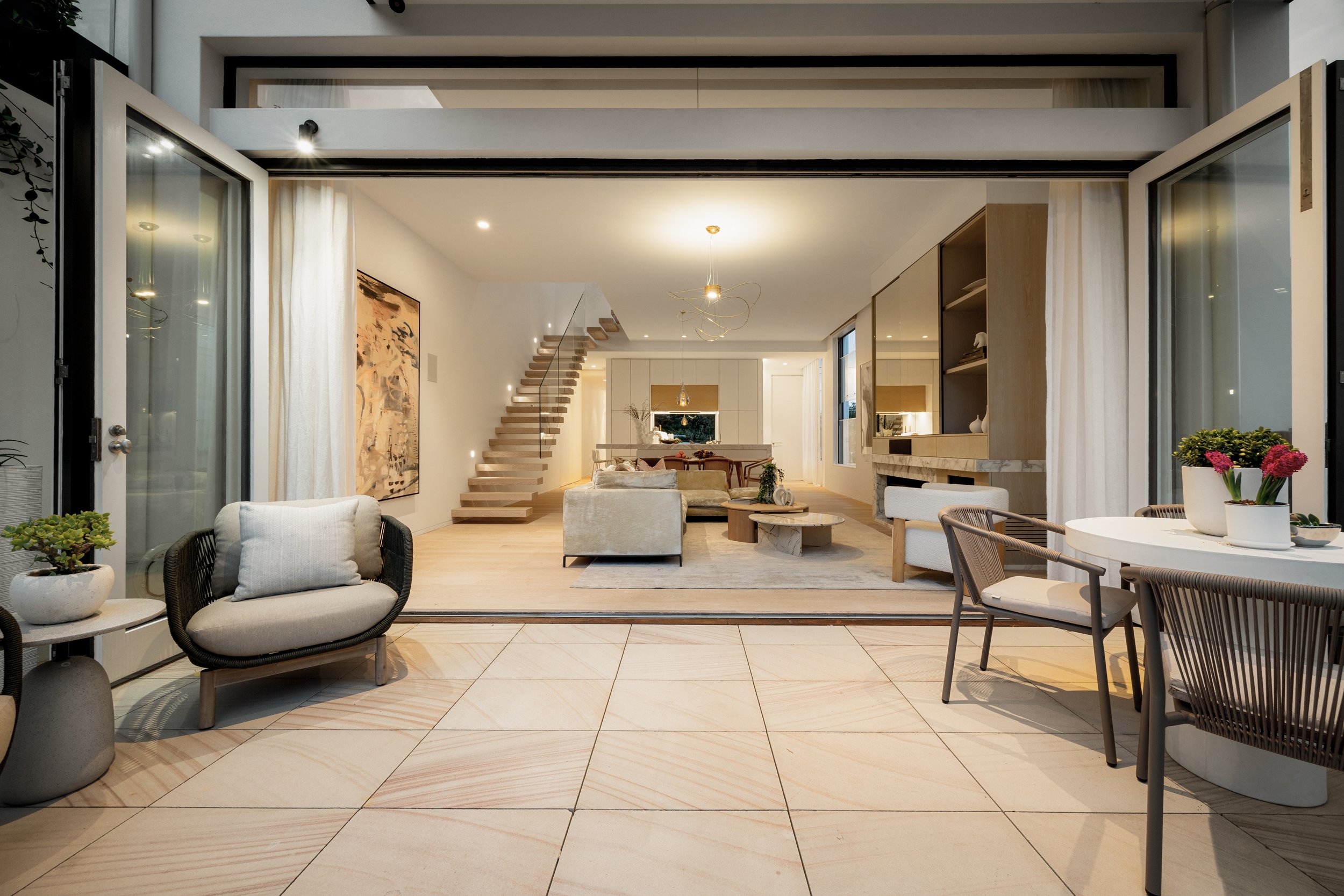 Open patio door leading to a modern, well-lit living room with a staircase, sitting area, artwork, and contemporary decor.