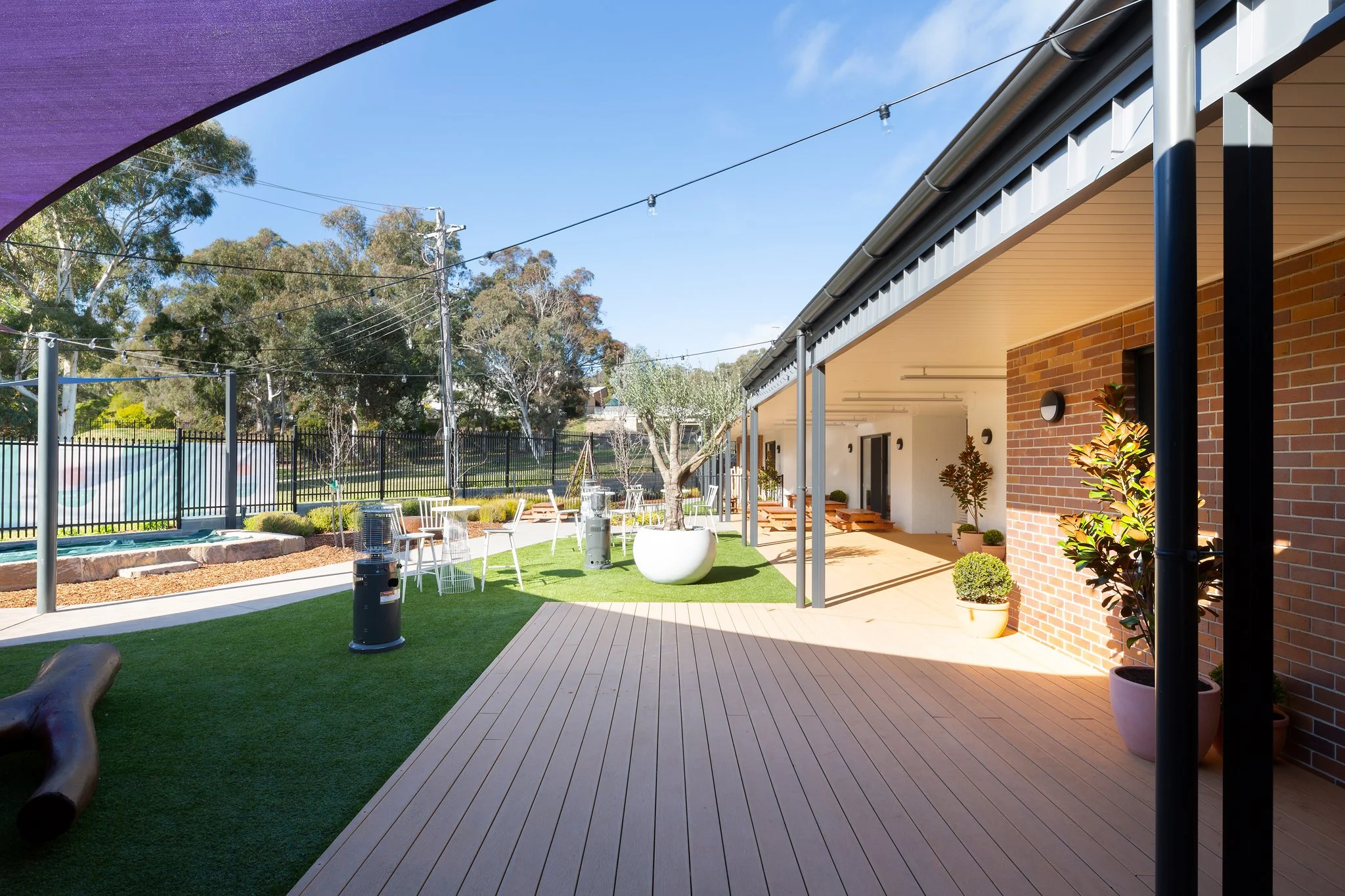 A backyard patio overlooking a fenced yard with trees, outdoor seating, potted plants, and a building with a brick wall and a covered porch area, under a clear blue sky.