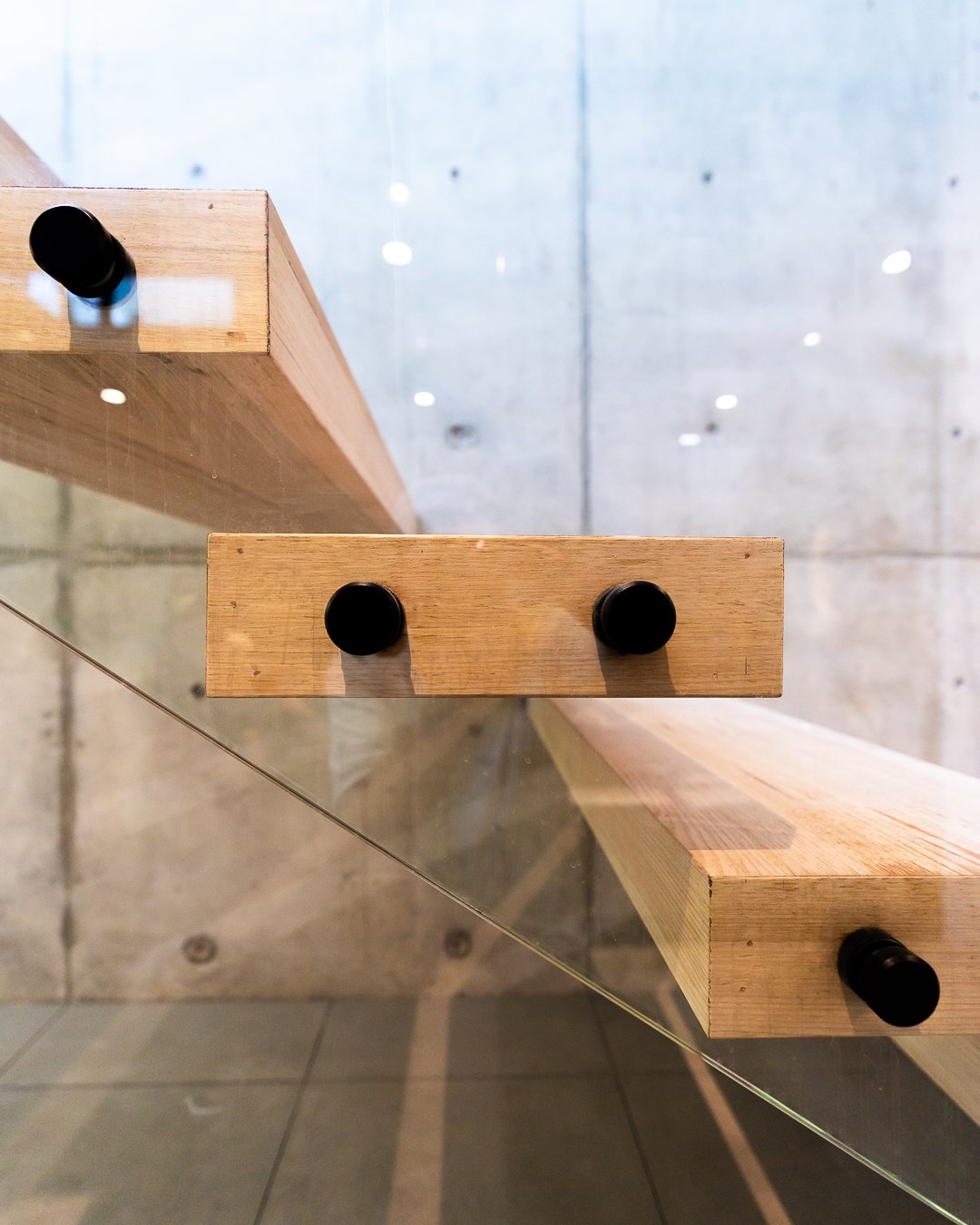 Close-up view of a floating wooden staircase with black rubber mounts, taken from below, with a glass panel and concrete wall in the background.