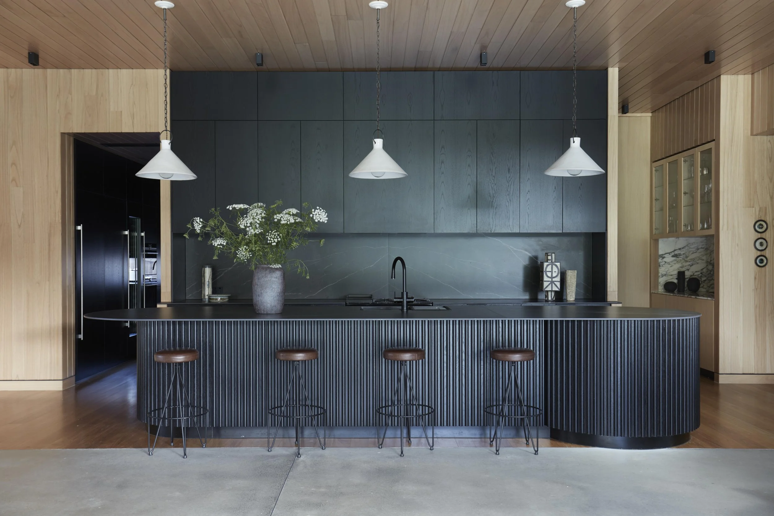 Modern kitchen with black cabinetry, wooden walls and ceiling, a black kitchen island with three stools, and a vase with white flowers on the island.