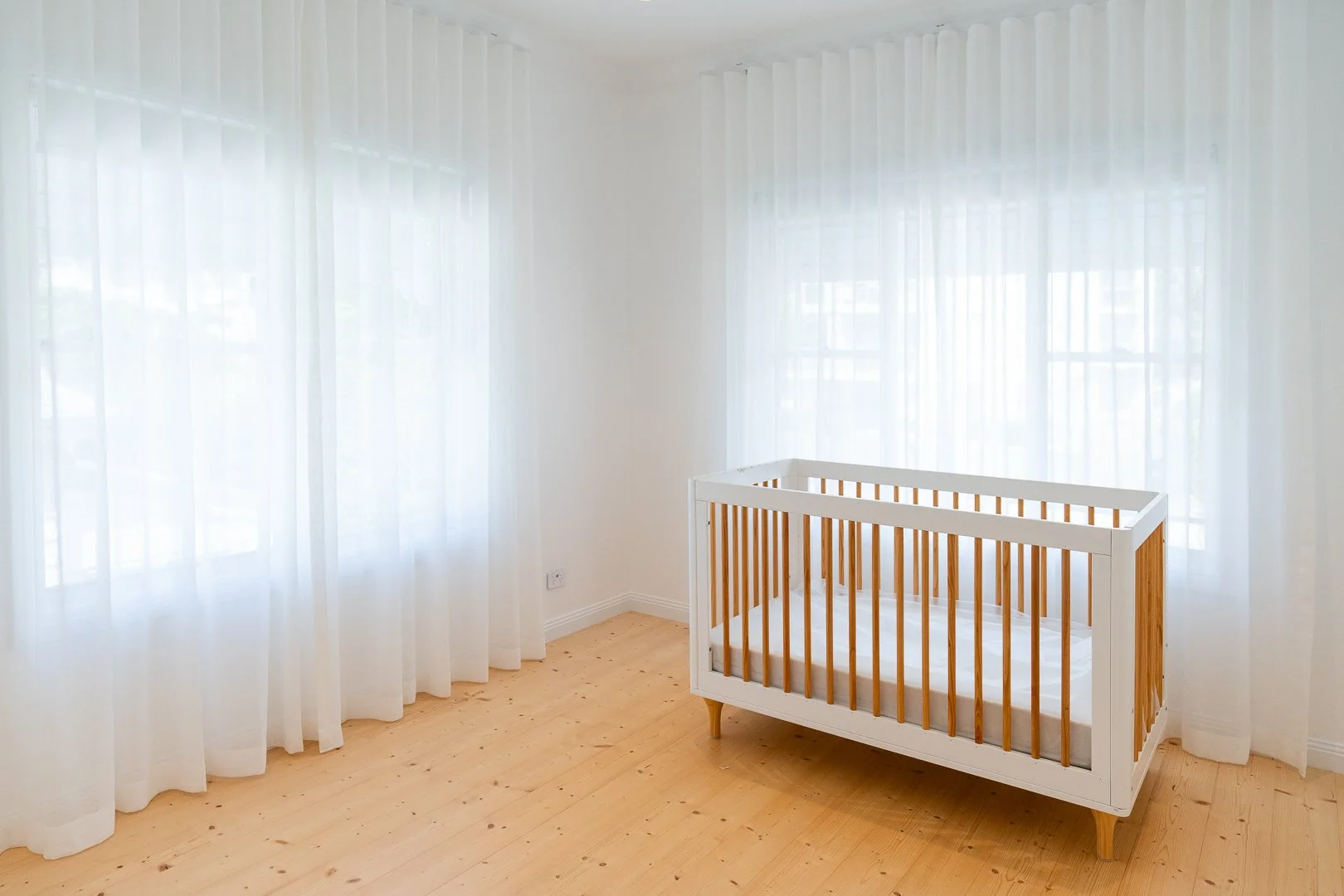 A nursery room with white curtains, light wooden flooring, and a white crib with wooden slats.