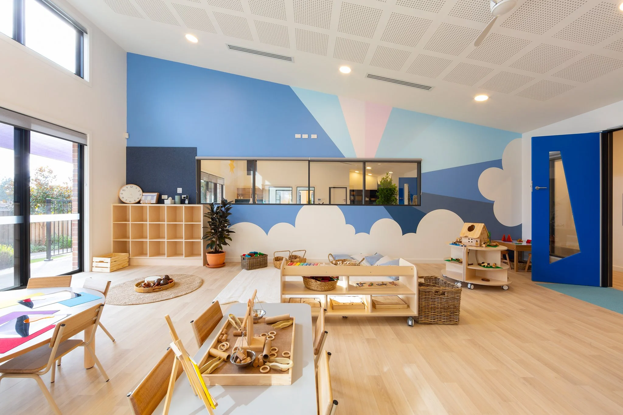 A brightly lit classroom with wood flooring, featuring a wall painted with clouds and pastel colors, and furniture such as small tables, chairs, shelves, and play areas for children.