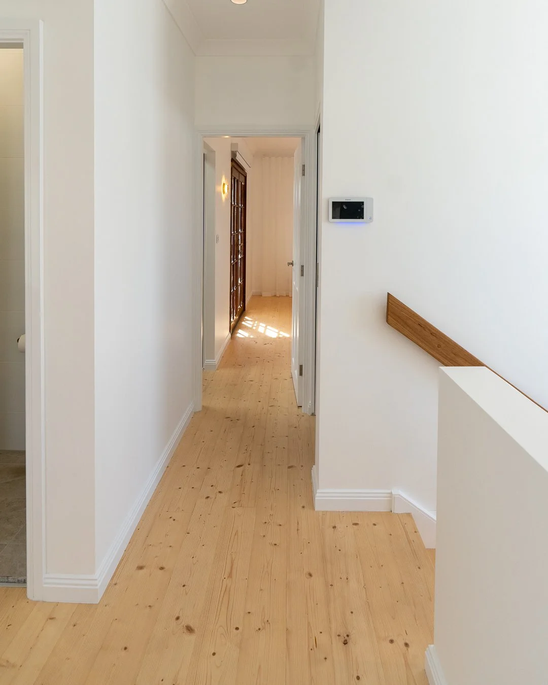 A hallway with light-colored wooden flooring and white walls, featuring a security or thermostat device mounted on the right wall and a wooden handrail on the right side.