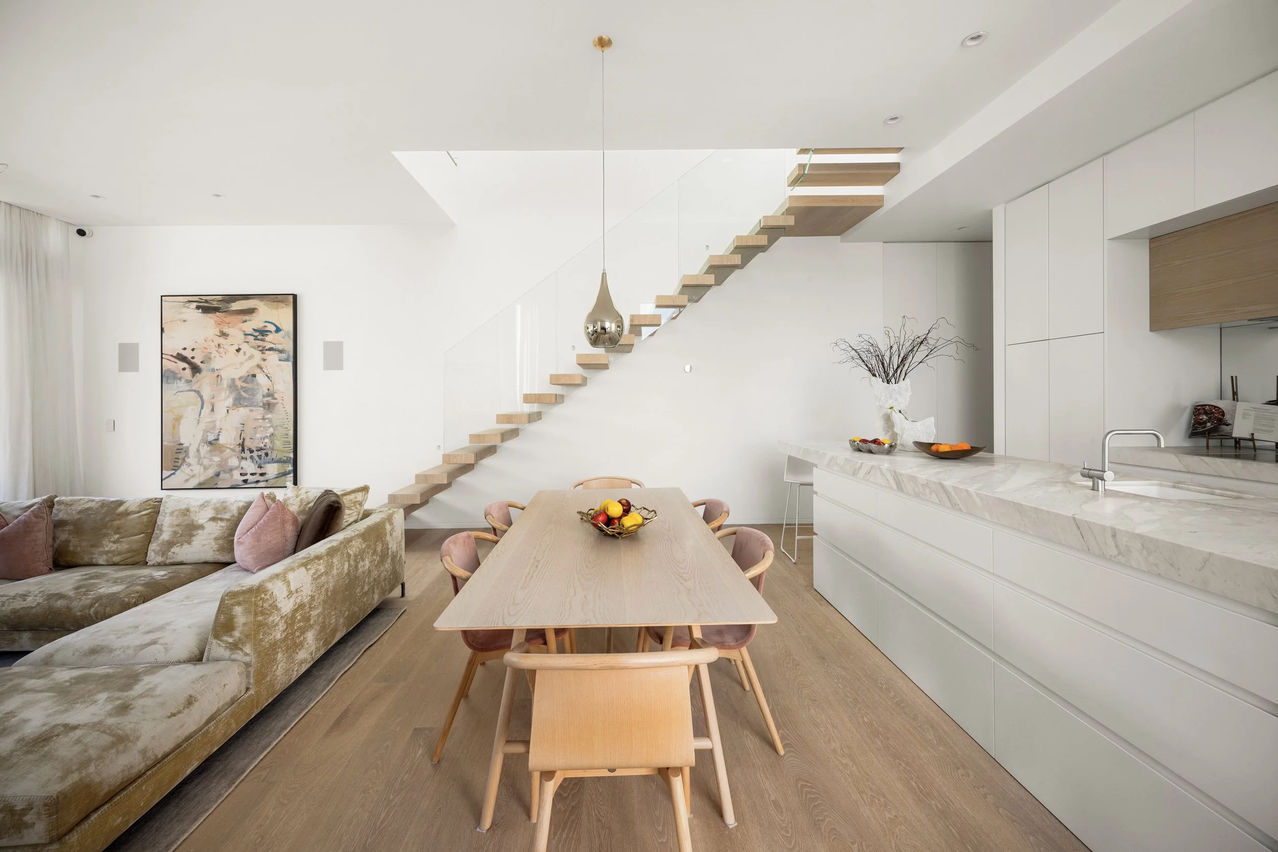 Modern living space with kitchen, dining area, staircase, beige sofa, abstract wall art, and minimal decor.