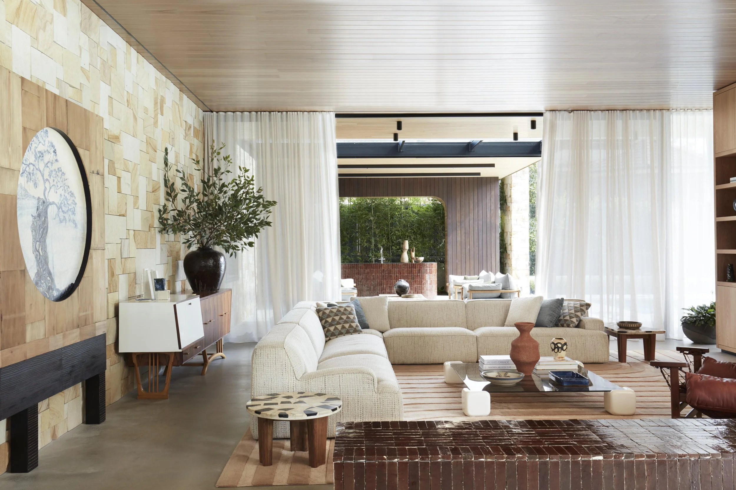 Bright living room with beige sectional sofa, wood coffee table, and decorative vases, lit by natural light with sheer curtains and a green outdoor view.
