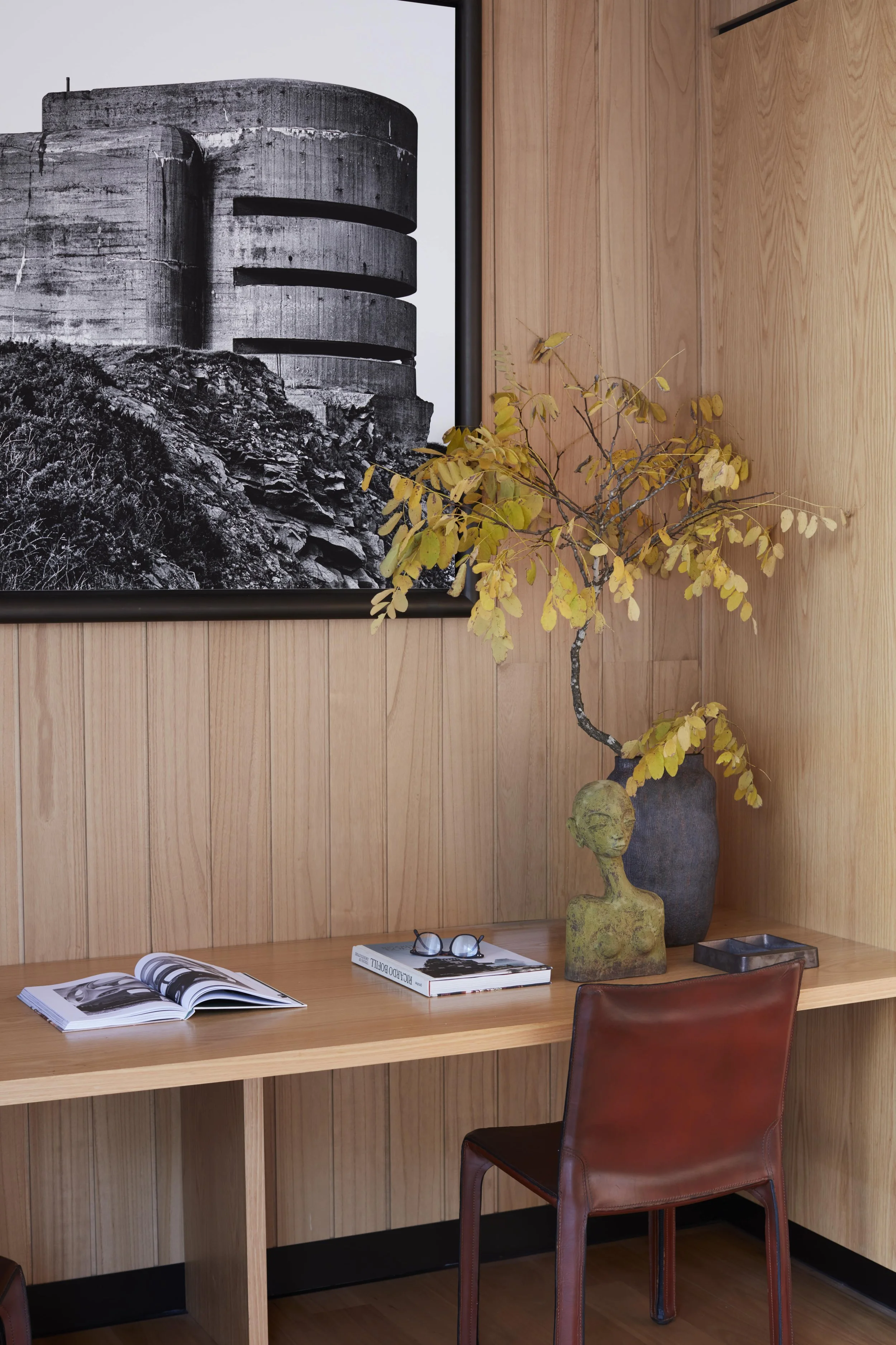 A wooden desk with an open magazine, a closed book, and a pair of glasses on top. Next to the desk are a tall ceramic vase with a yellow-green leafy branch and a smaller ceramic sculpture resembling a human figure. Behind the desk, a black and white 