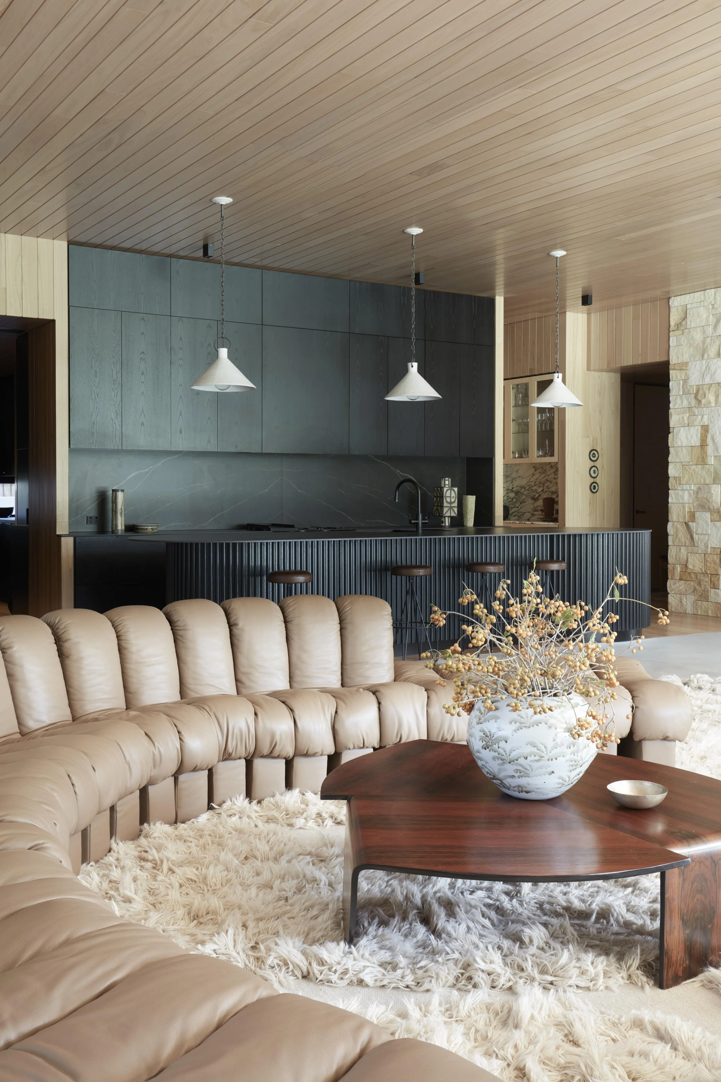 Living room with beige tufted sectional sofa, shaggy white rug, dark wood coffee table with a large white vase with dried flowers, and a beige curtain window with wooden accents in the background.