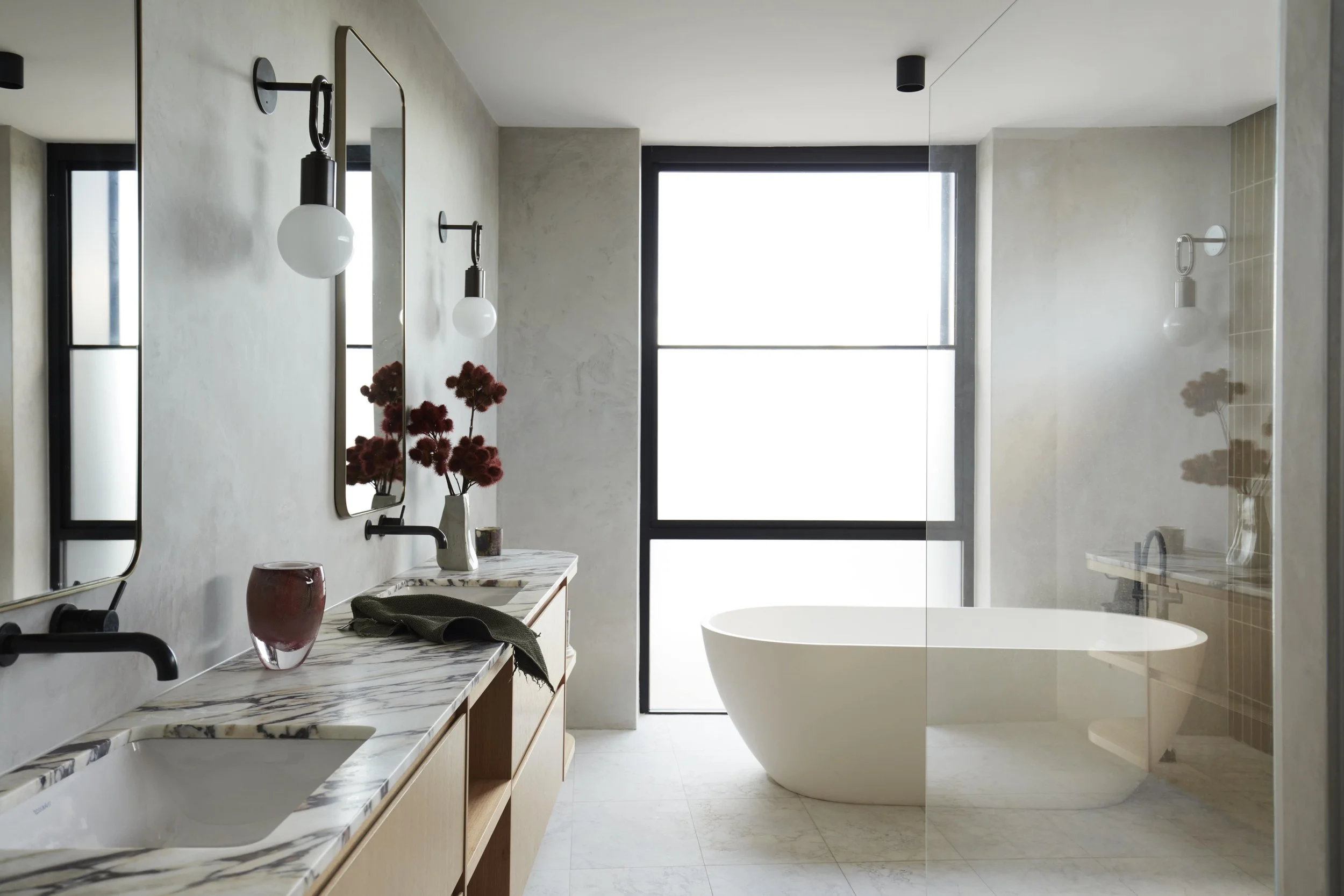 Modern bathroom with a freestanding bathtub, large window, marble countertop with double sinks, mirror, wall-mounted lamps, and decorative flower vase.