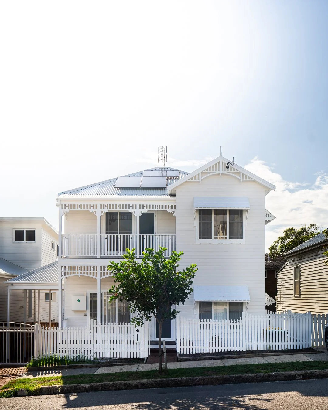 White two-story house with decorative trim, a front porch with railing, a small tree in front, surrounded by a white picket fence, under a bright sky.