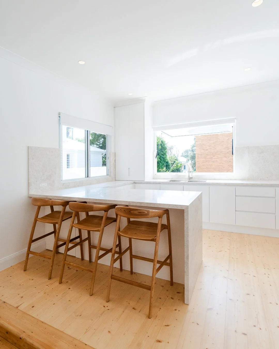 Bright, modern kitchen with white cabinets, marble countertop island, and three wooden bar stools. Large windows provide natural light and a view of outdoor greenery.