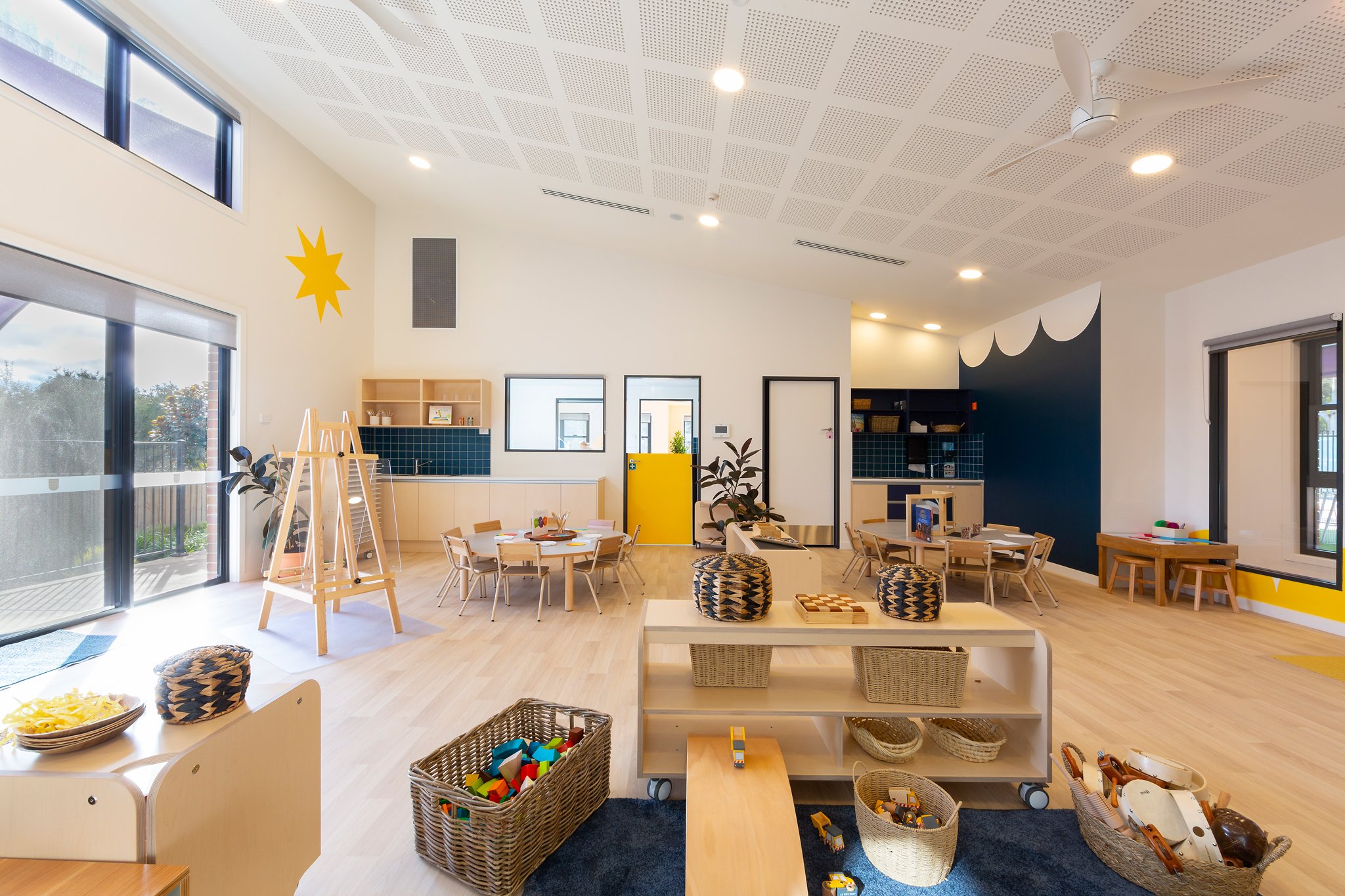 Interior of a preschool or daycare room with natural light, wooden furniture, and shelves filled with toys and books. The room features a blue and yellow color scheme, with tables and chairs for children, and various play areas with building blocks a