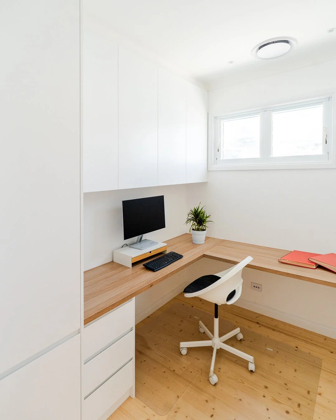Minimalist home office with white walls, wooden desk, white rolling chair, small computer monitor, keyboard, a plant, and some closed folders on the desk, in front of a window.