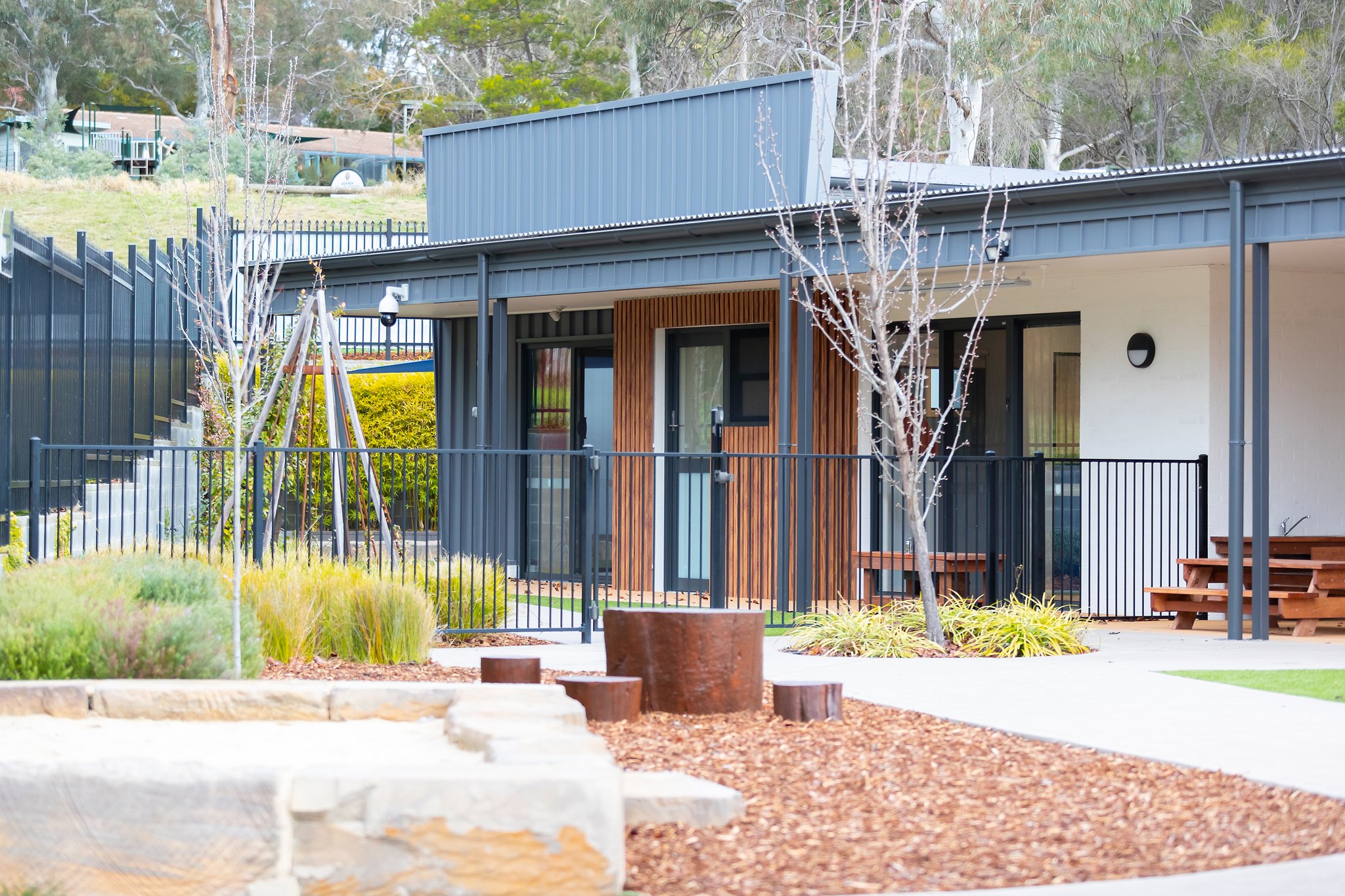 Modern building with metal siding, wooden accents, glass doors, and a fenced outdoor area, surrounded by trees and landscaping.