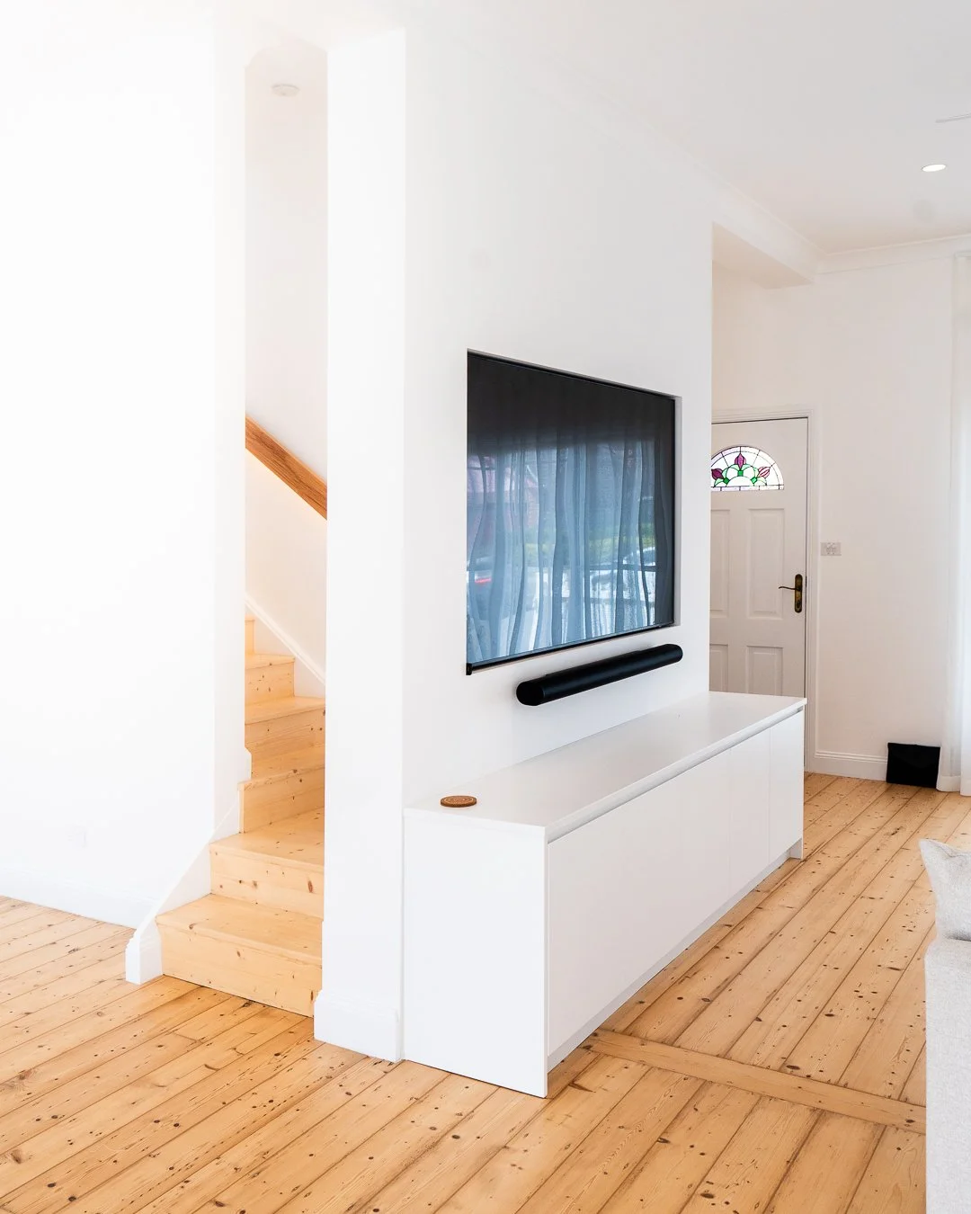Living room with white walls, wooden flooring, a mounted TV with a soundbar, white cabinet, and a door with stained glass window.