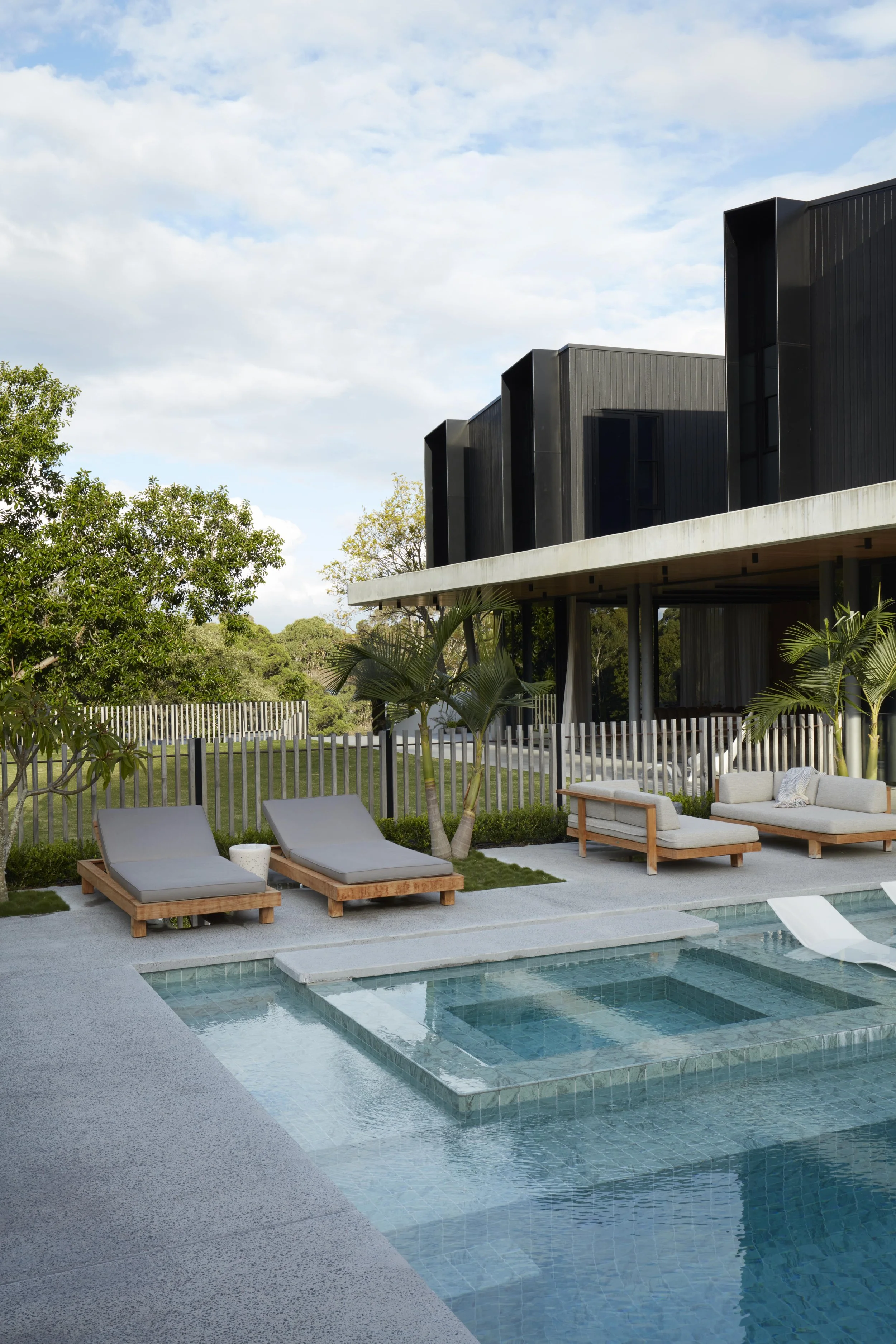 Modern house with black exterior on a raised foundation, outdoors with a pool, lounge chairs, potted plants, trees, and a fence under a partly cloudy sky.