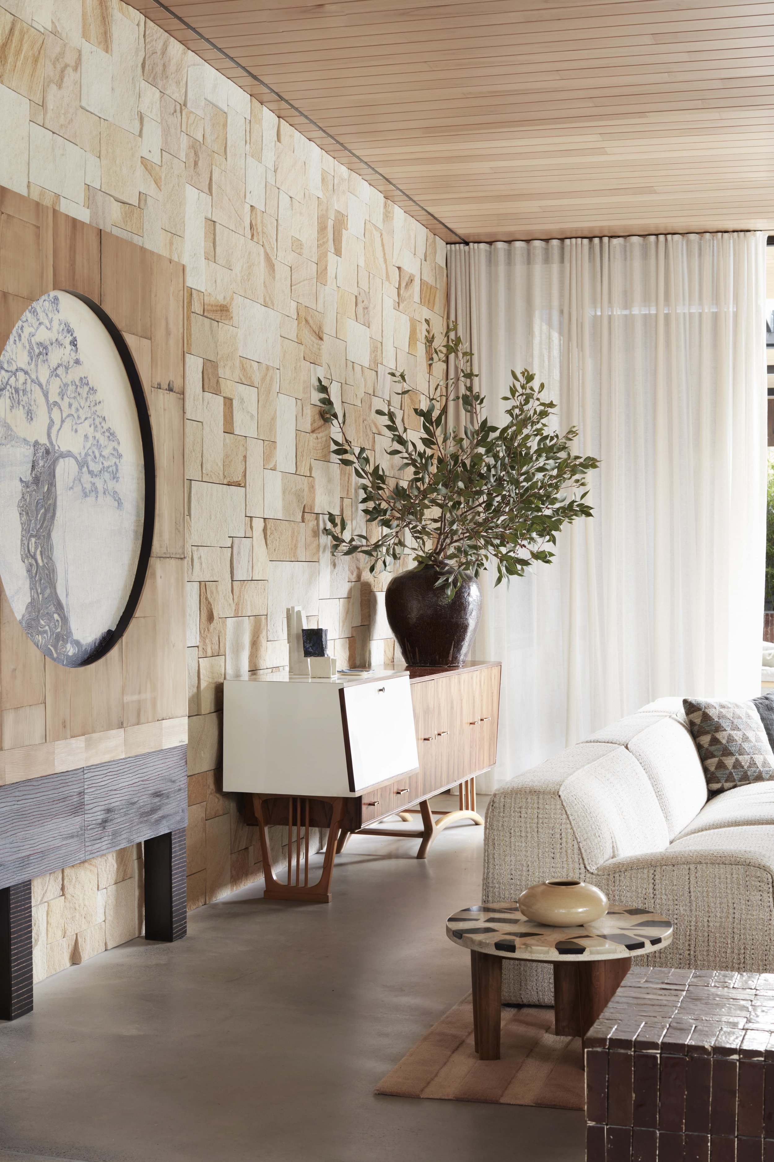 Living room with stone accent wall, wooden ceiling, cream-colored sofa with throw pillows, large window with white curtains, side table with two decorative vases, and a wooden standing cabinet with a large plant in a dark vase on top.