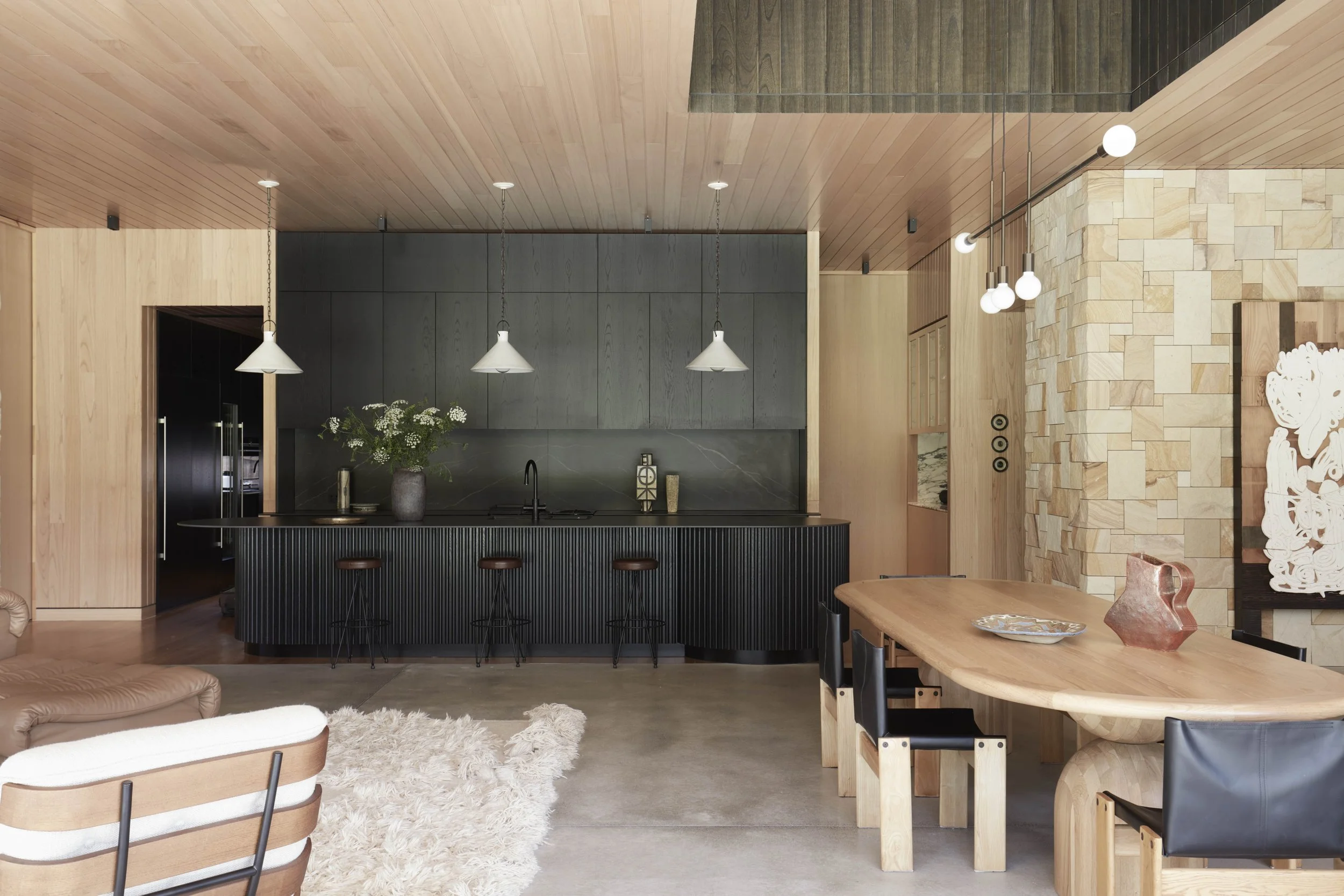 Modern kitchen and dining area with wooden ceiling and stone wall, featuring pendant and globe lighting, a black kitchen island, and a dining table with chairs.