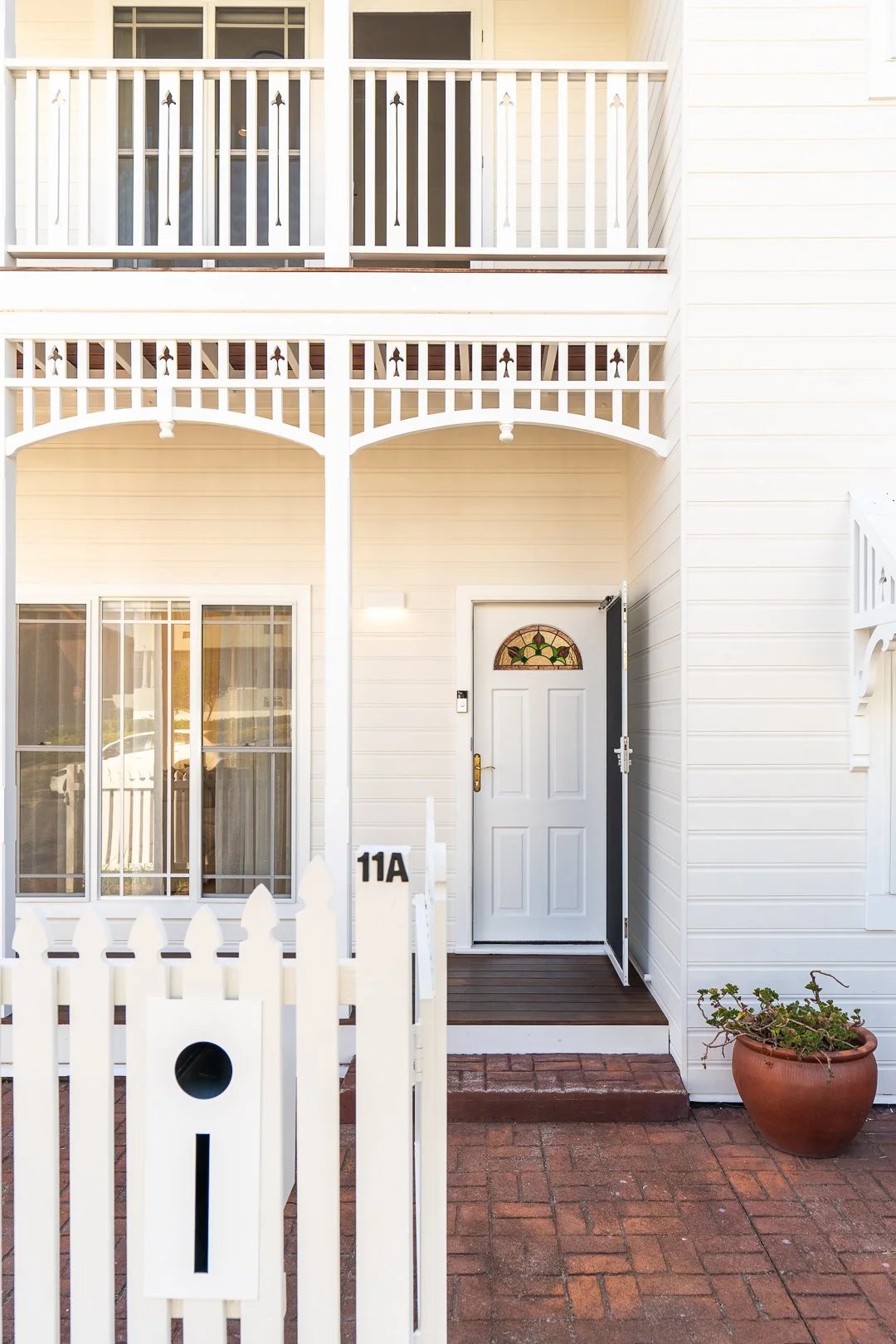 Front view of a white house with a small porch, decorative balcony, white picket fence, and a potted plant outside the front door.