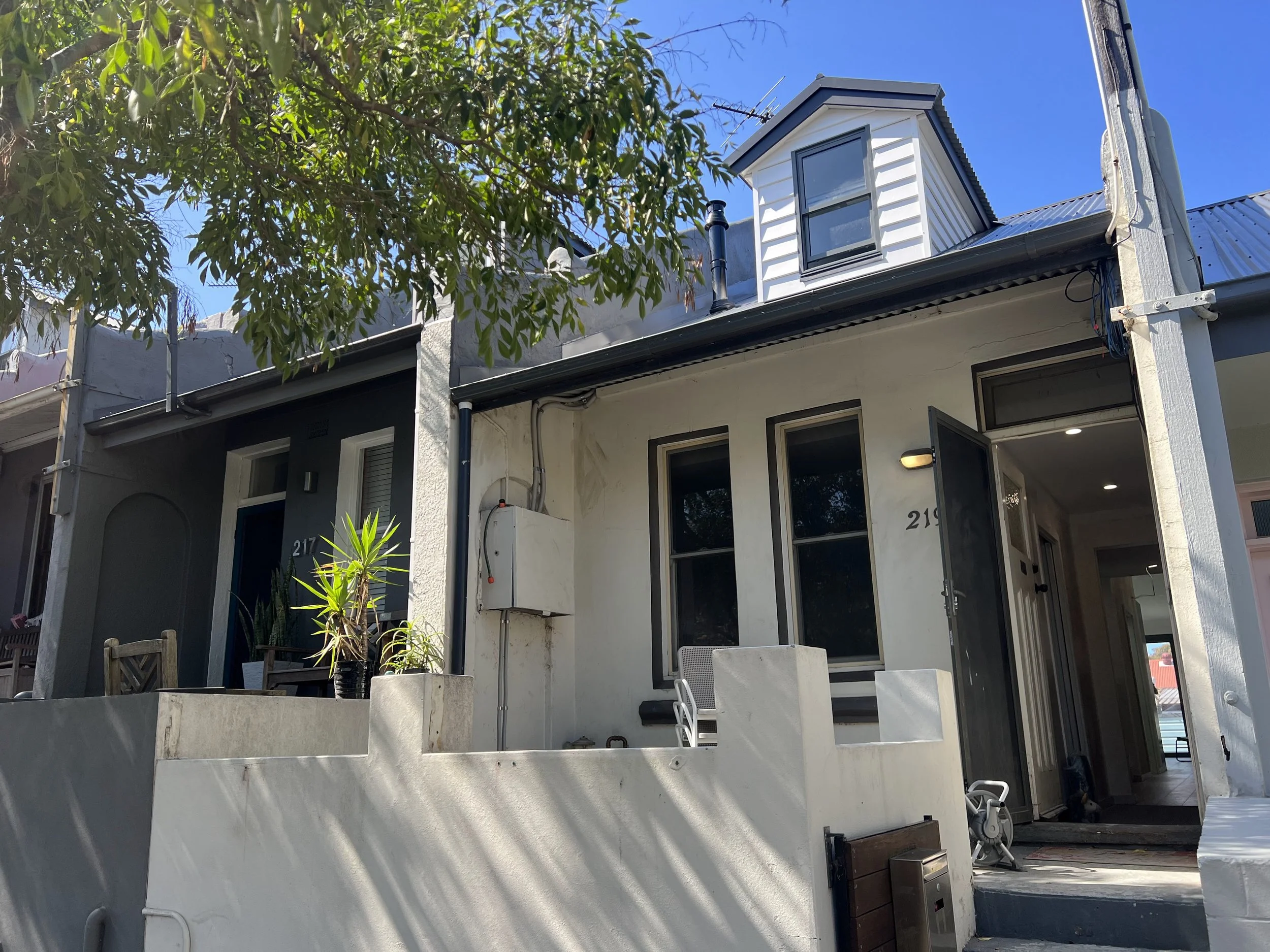 Exterior of a modern house with a dormer window on the roof, a small porch area, some greenery, and construction or renovation materials visible.