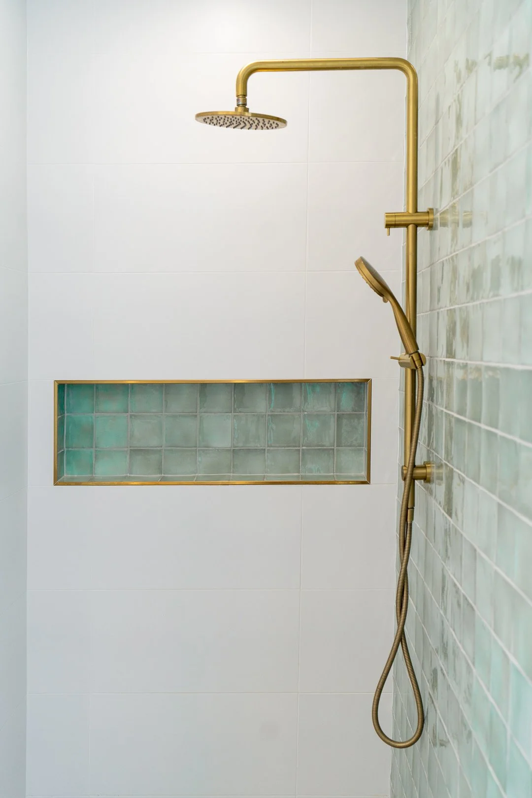 Modern shower with a brass showerhead and a handheld shower wand, against a white tiled wall with a horizontal niche containing green glass tiles.