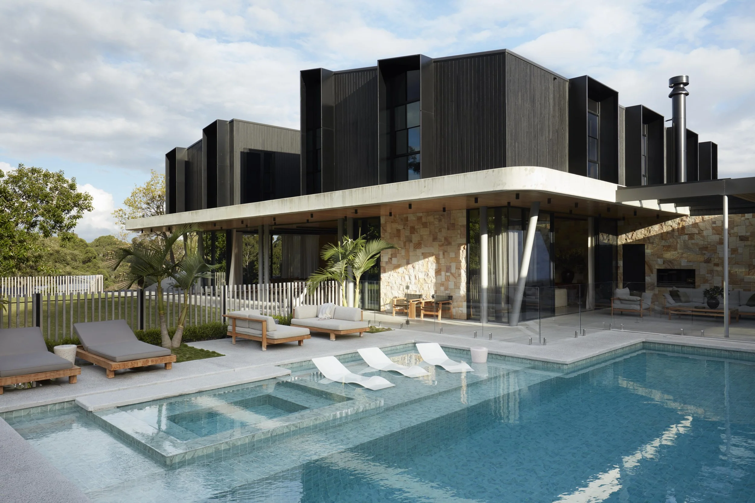 Modern house with a pool and outdoor seating area, surrounded by trees and fenced yard, under a partly cloudy sky.