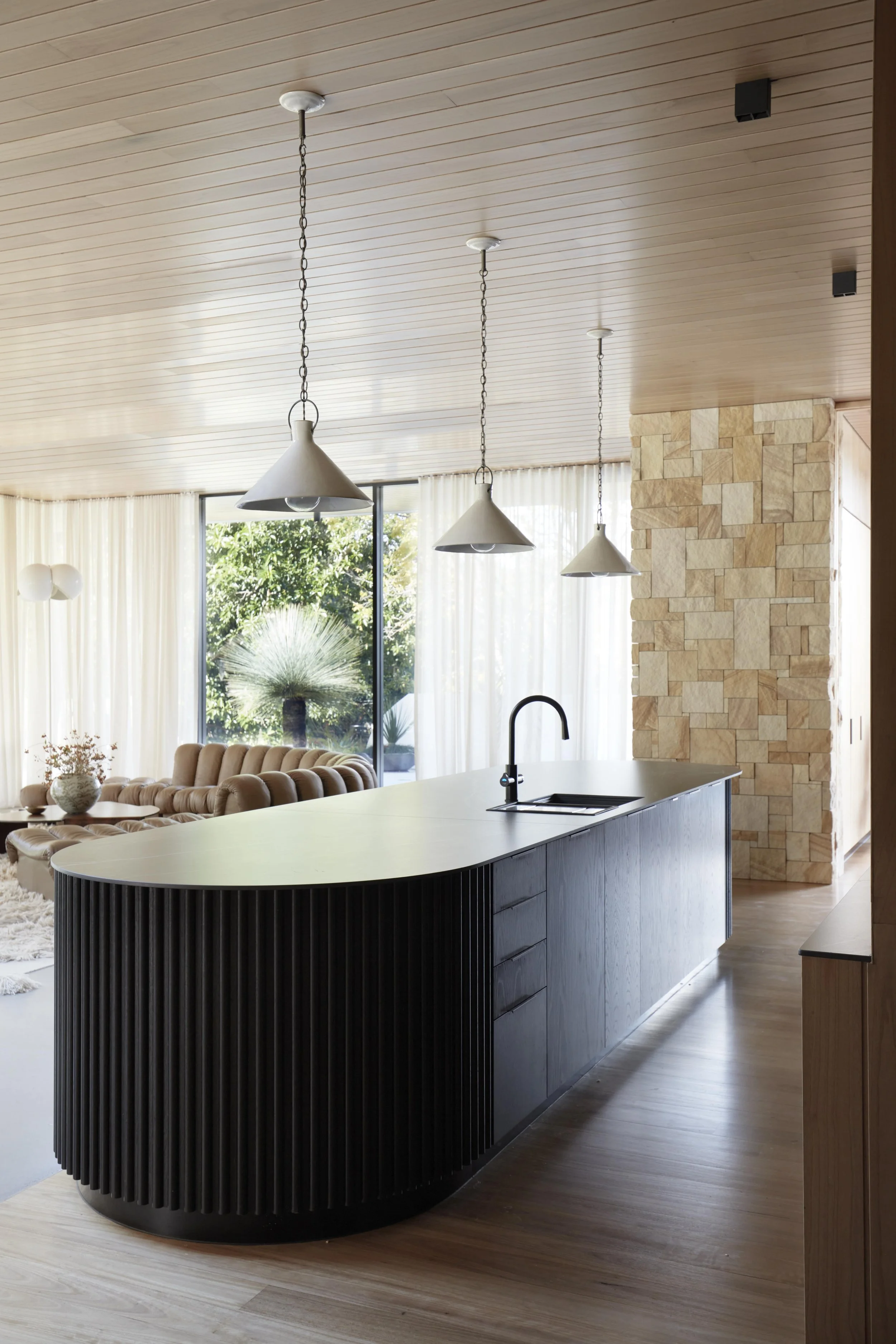 Modern kitchen with black curved island, beige stone wall, and three white pendant lights hanging from a wooden ceiling. Large window with curtains overlooks greenery outside.