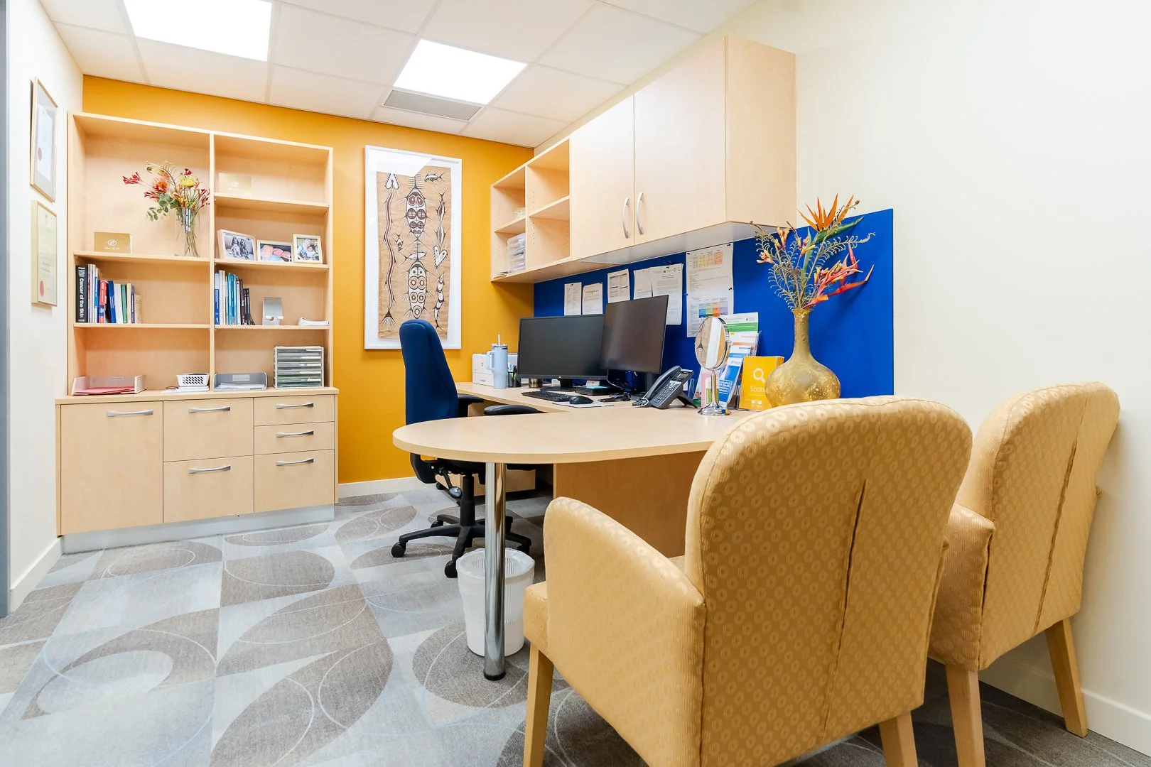 Office with two computers on a desk, three yellow chairs, a bookshelf, and colorful artwork on the wall.