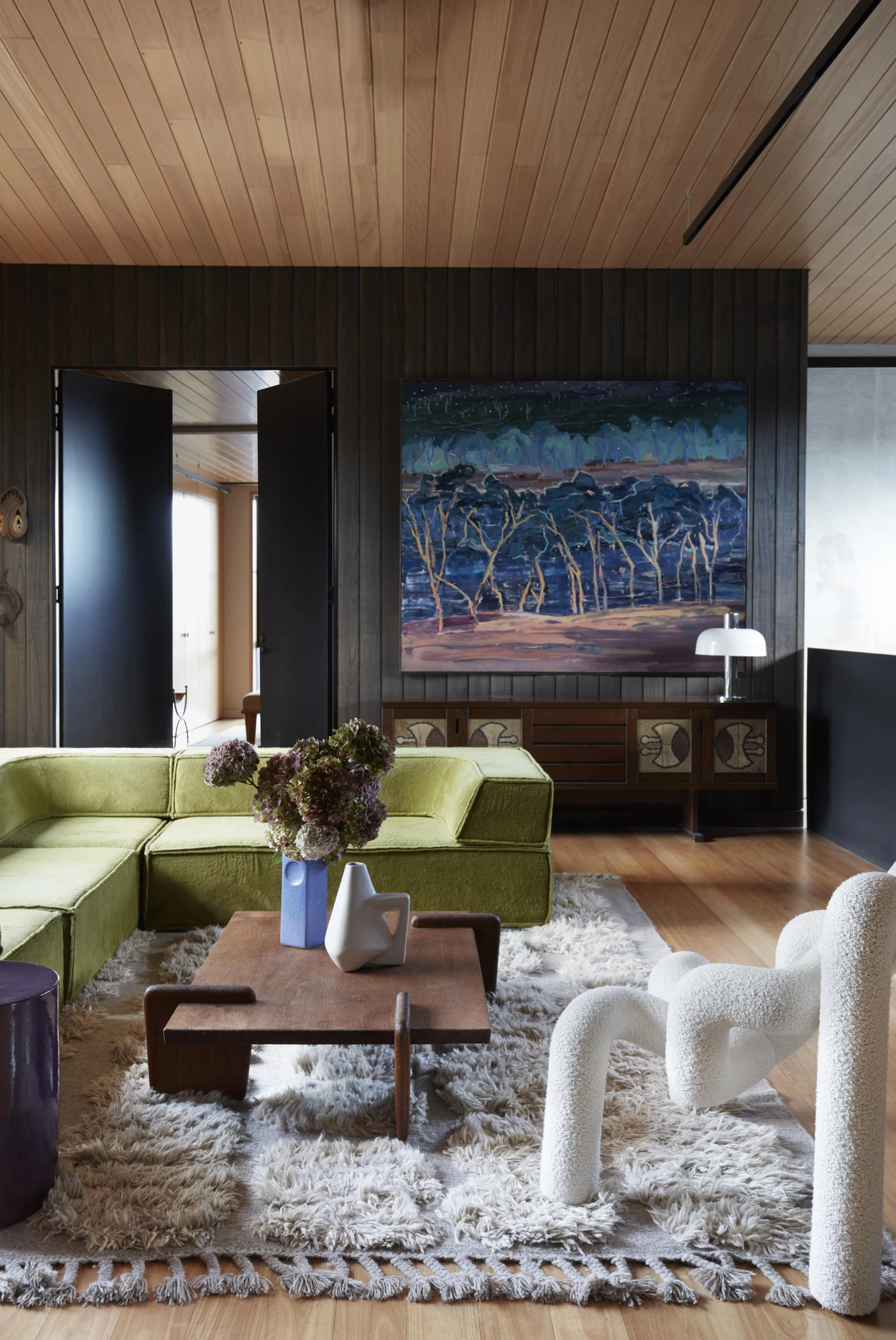 Interior of a modern living room with a vintage-inspired style. Features include a bright green velvet sofa, a wooden coffee table with vases, a white textured armchair, a large abstract painting of a landscape, a shaggy cream rug, and a dark wood wa