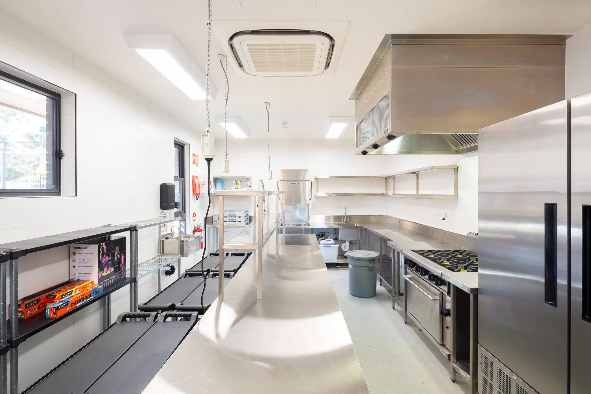 Clean commercial kitchen with stainless steel appliances, countertops, shelves, a stove with burners, and a window on the left wall, well-lit by fluorescent lights.