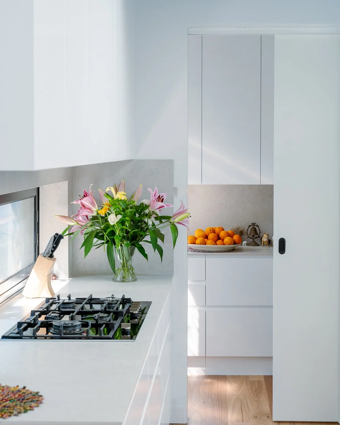 A modern kitchen with white cabinets and countertops, a Built-in gas stove, a vase of pink lilies and yellow flowers, and a cabinet with a bowl of oranges.