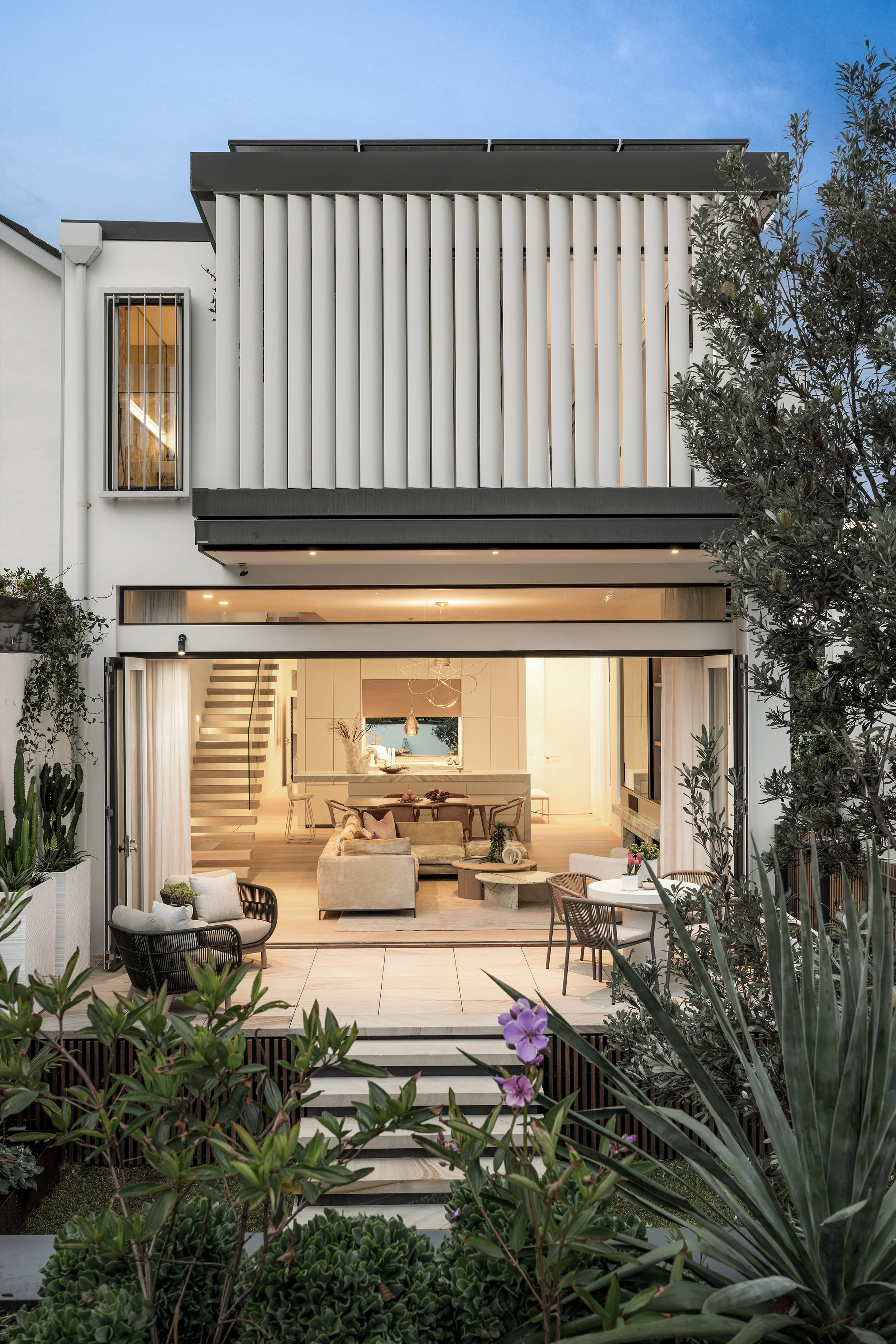 Modern house with open living space, large glass doors, and an outdoor patio with garden plants and seating, illuminated in the evening.
