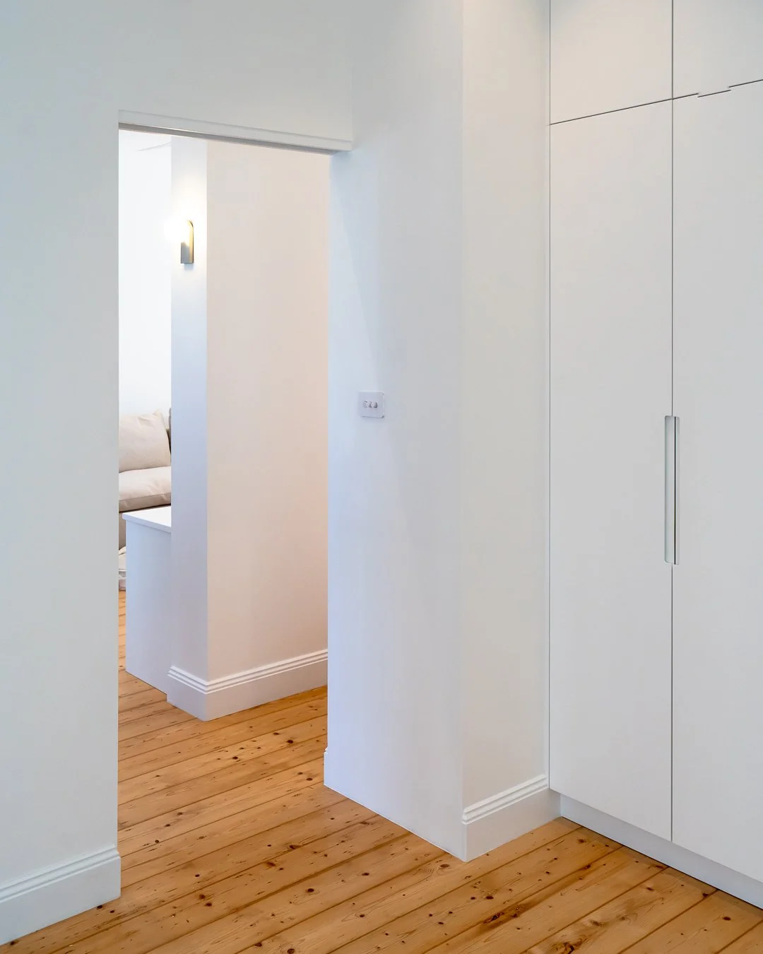Interior view of a white room with hardwood flooring, a built-in storage cabinet, and a doorway leading to a bedroom with a beige bed and a wall-mounted lamp.