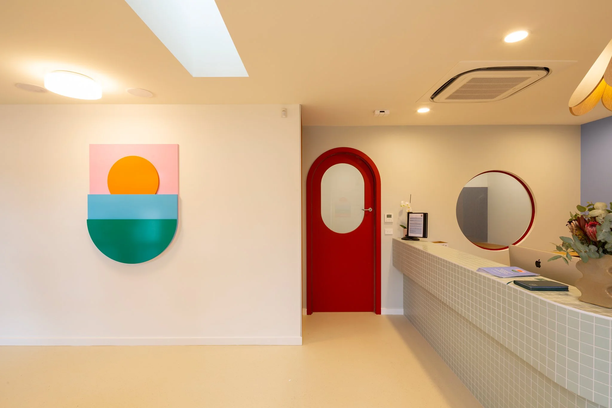 Interior of a modern room with a colorful abstract wall art, a red door with an oval window, a reception desk with a mirror and a computer, and ceiling lights.