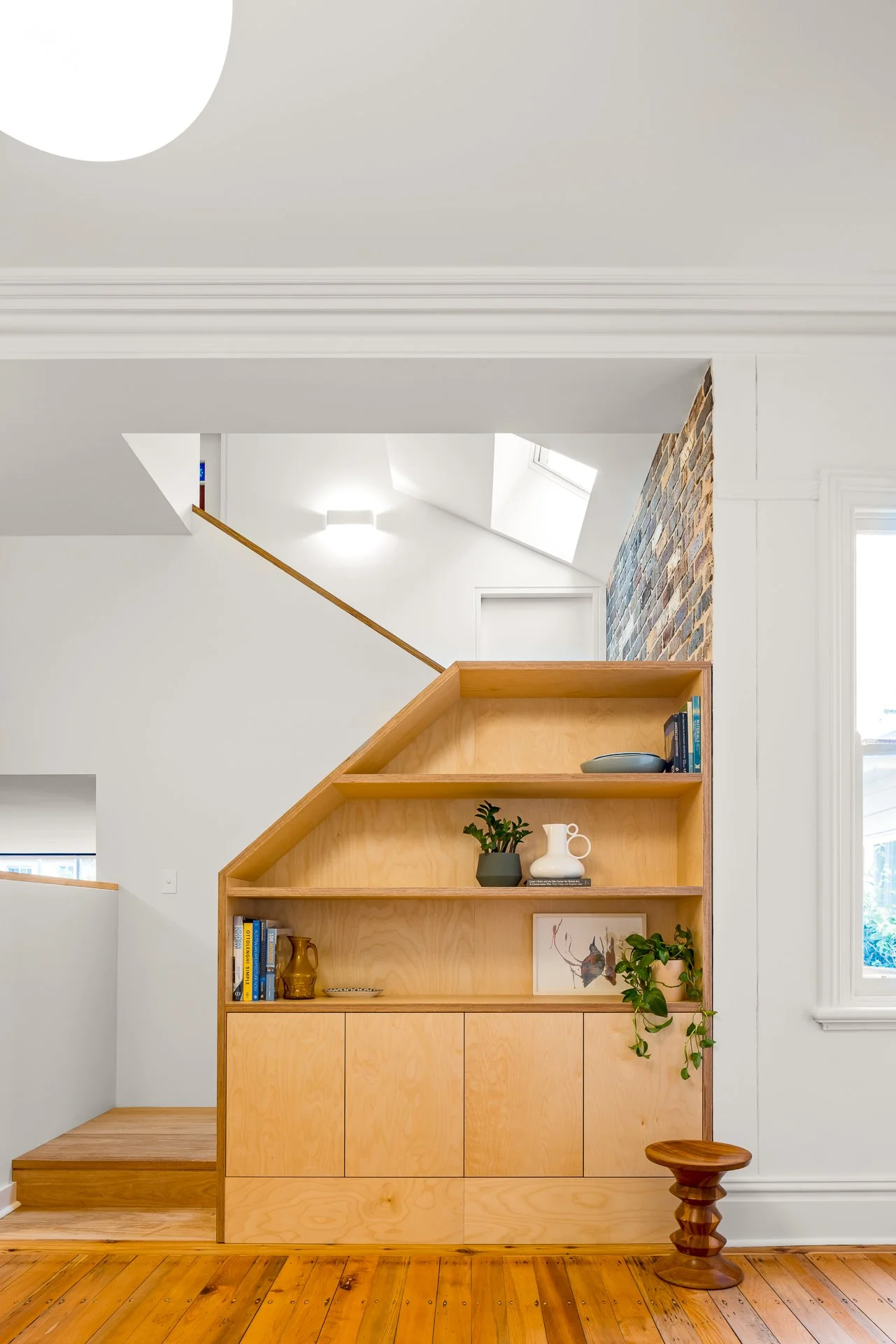 Interior of a modern home with a wooden bookshelf, decorative objects, plants, a staircase, a brick accent wall, and natural light coming through windows.