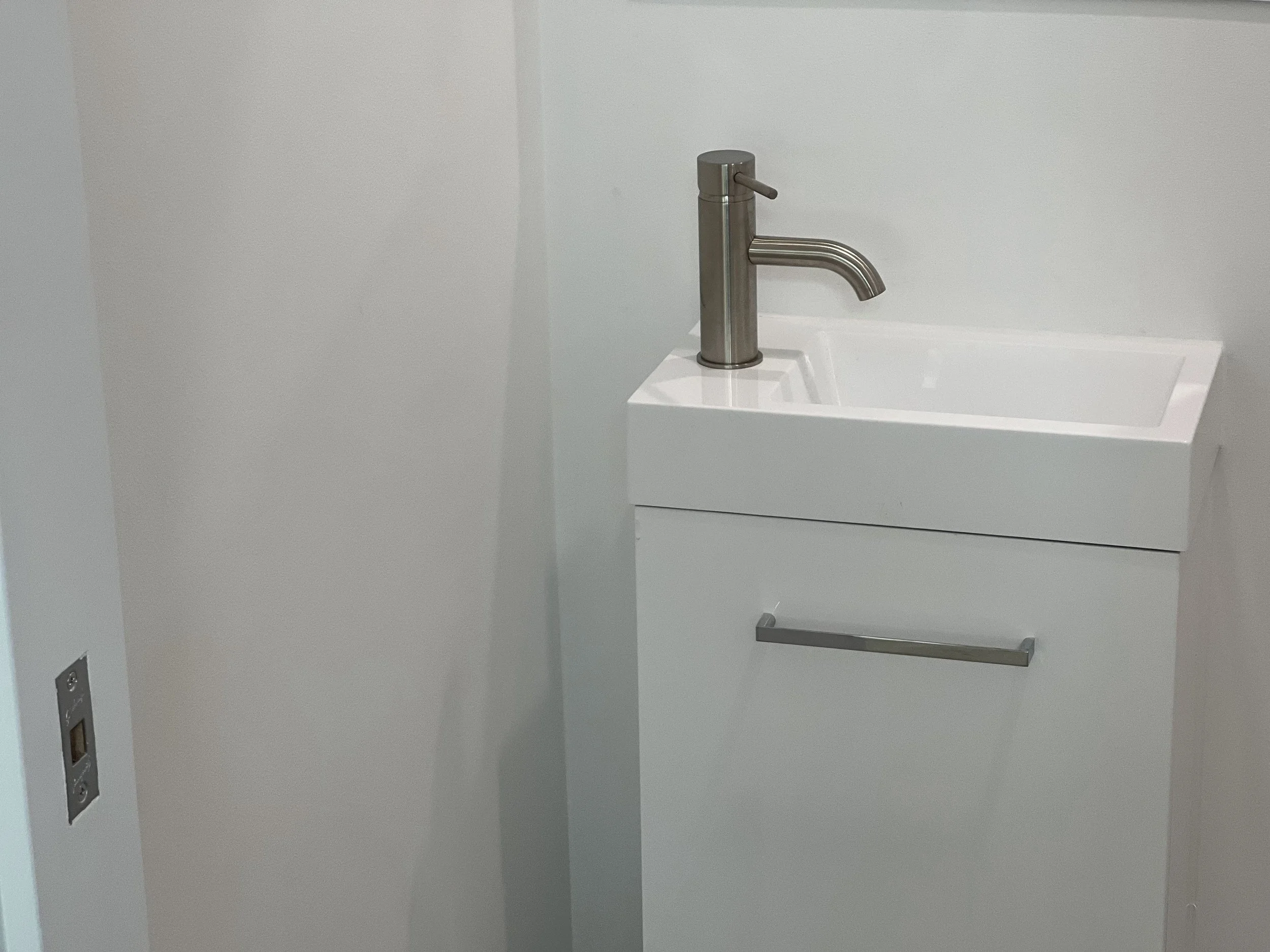 A modern, stainless steel faucet installed on a small white sink in a minimalist bathroom or utility area with plain white walls.