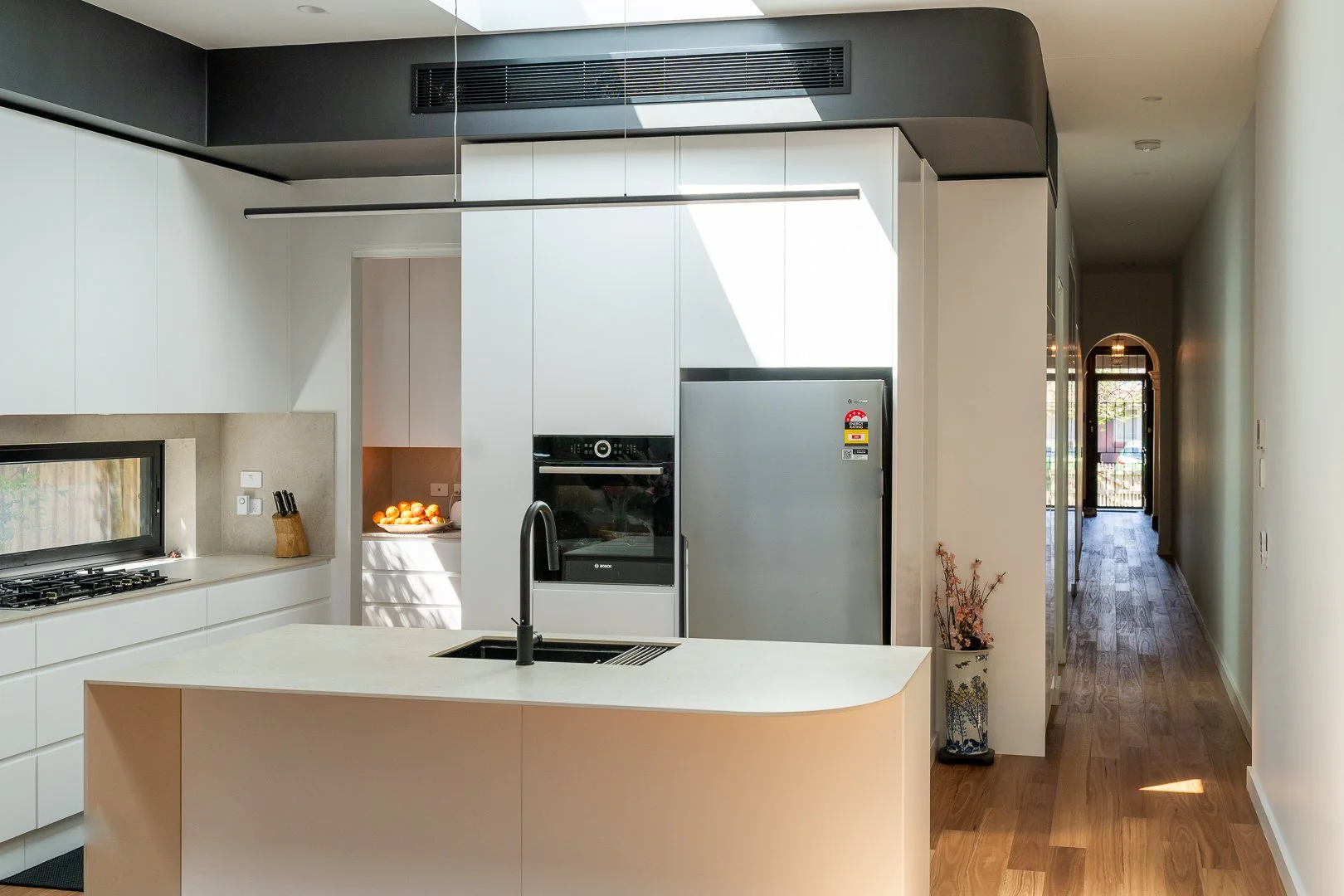 Modern kitchen with white cabinetry, black accents, and a kitchen island with a sink, stainless steel refrigerator, built-in oven, and hallway leading to another room with wooden flooring.