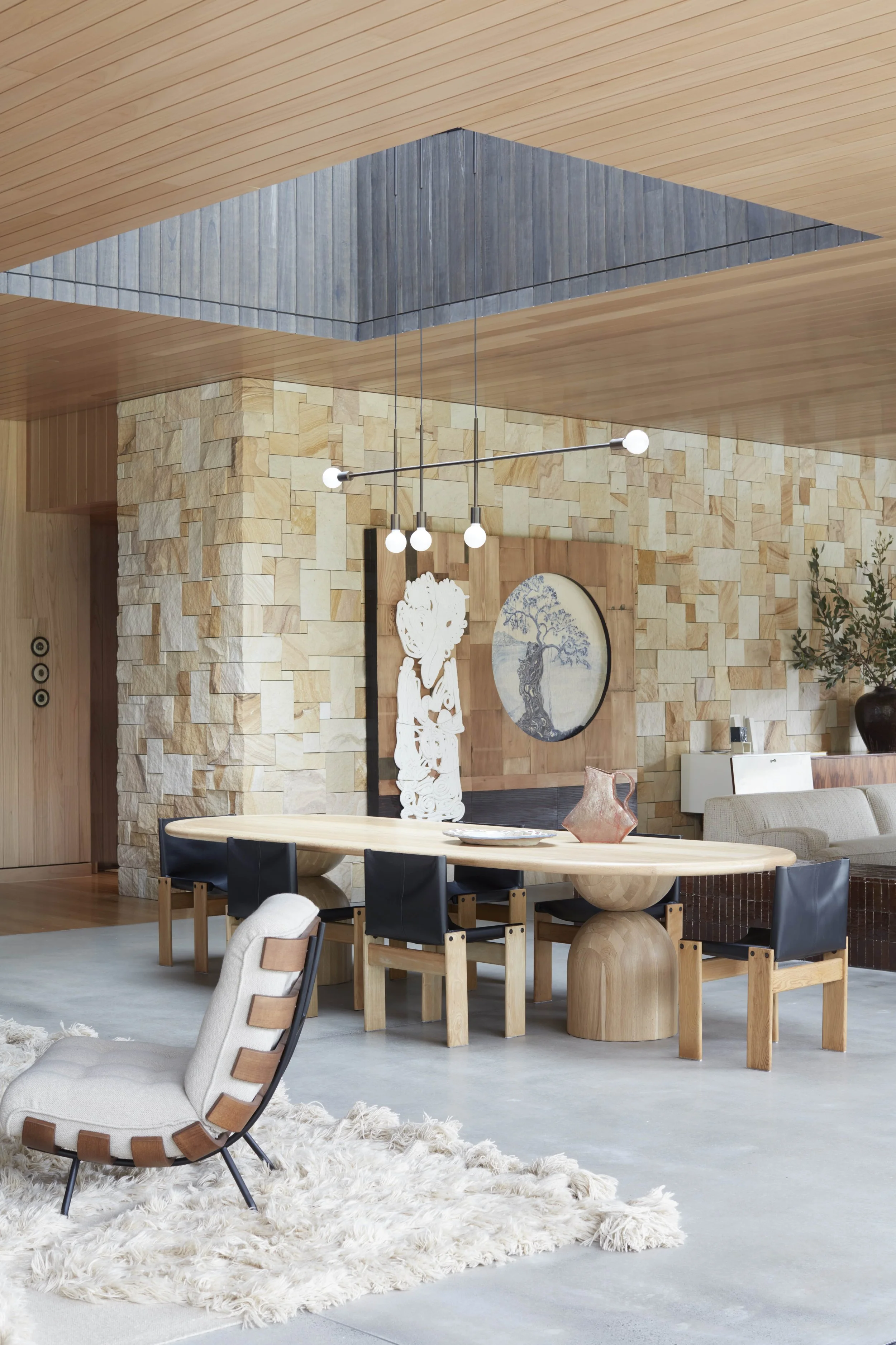 Modern interior living room with wood and stone walls, a long wooden dining table, black chairs, decorative art, and a shaggy white rug.