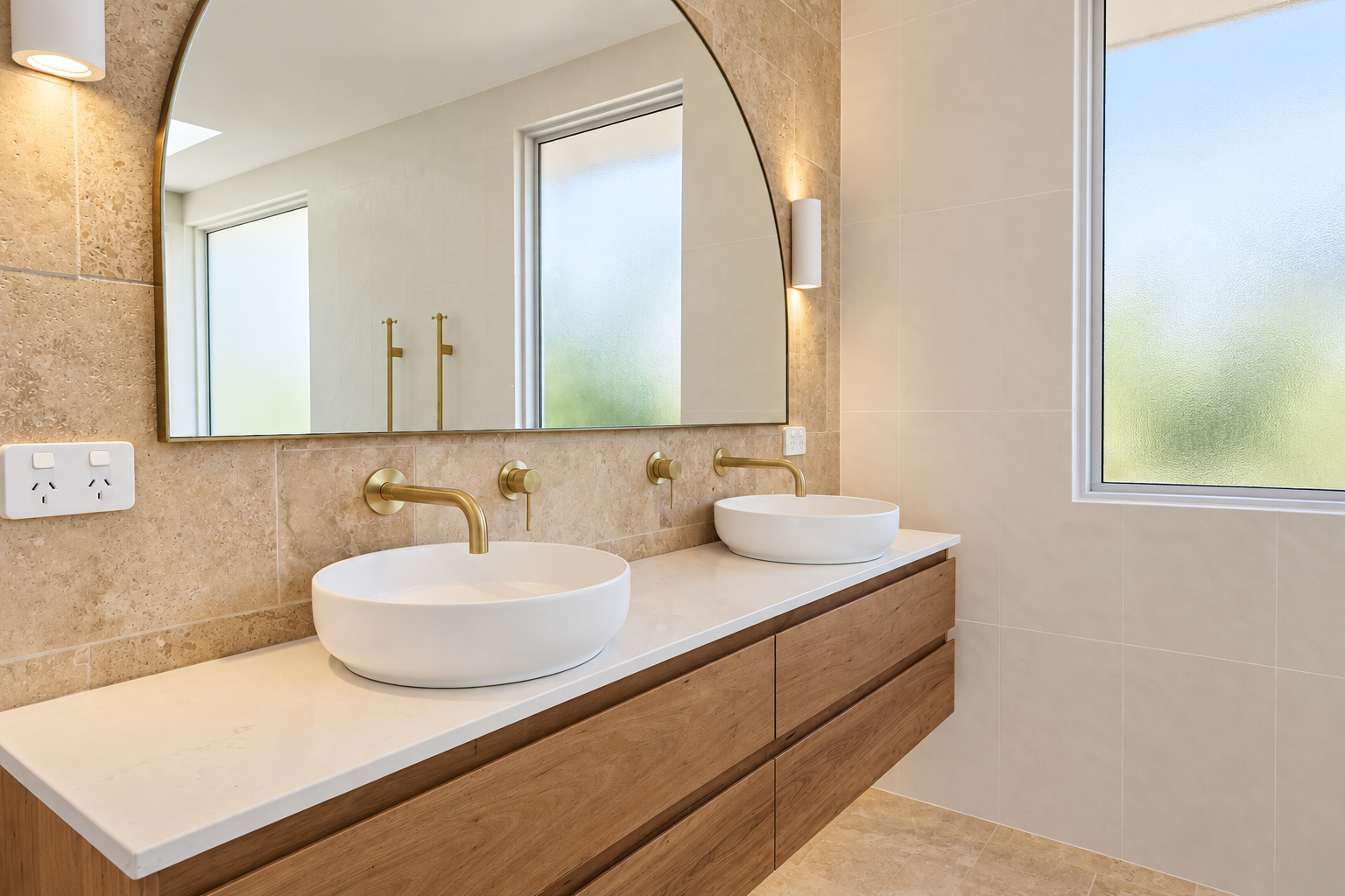 Modern bathroom with double white vessel sinks, gold fixtures, large wall mirror, and natural light from frosted window.