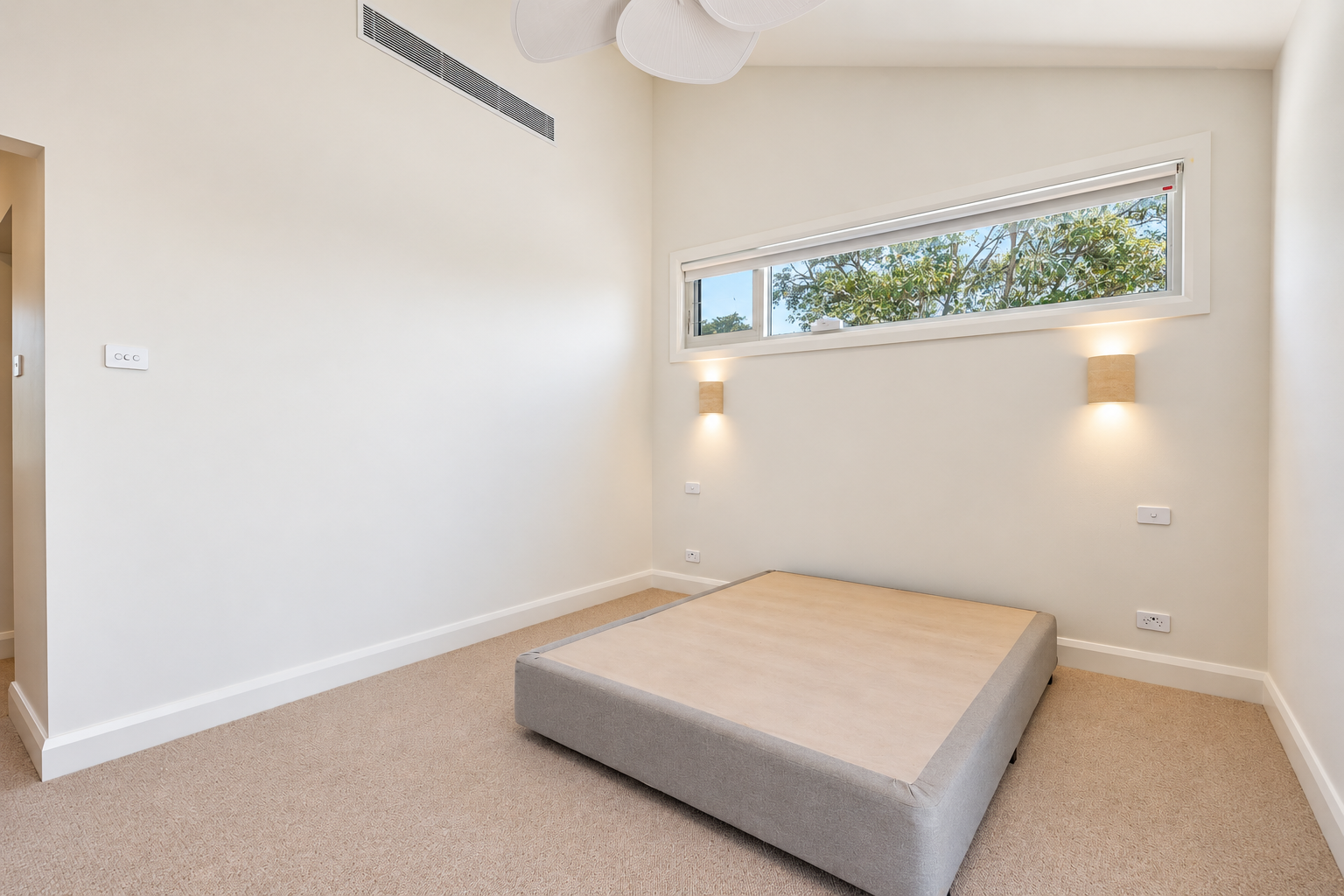 Empty room with a mattress on the floor, white walls, a large rectangular window, beige carpet, and wall-mounted lamps.