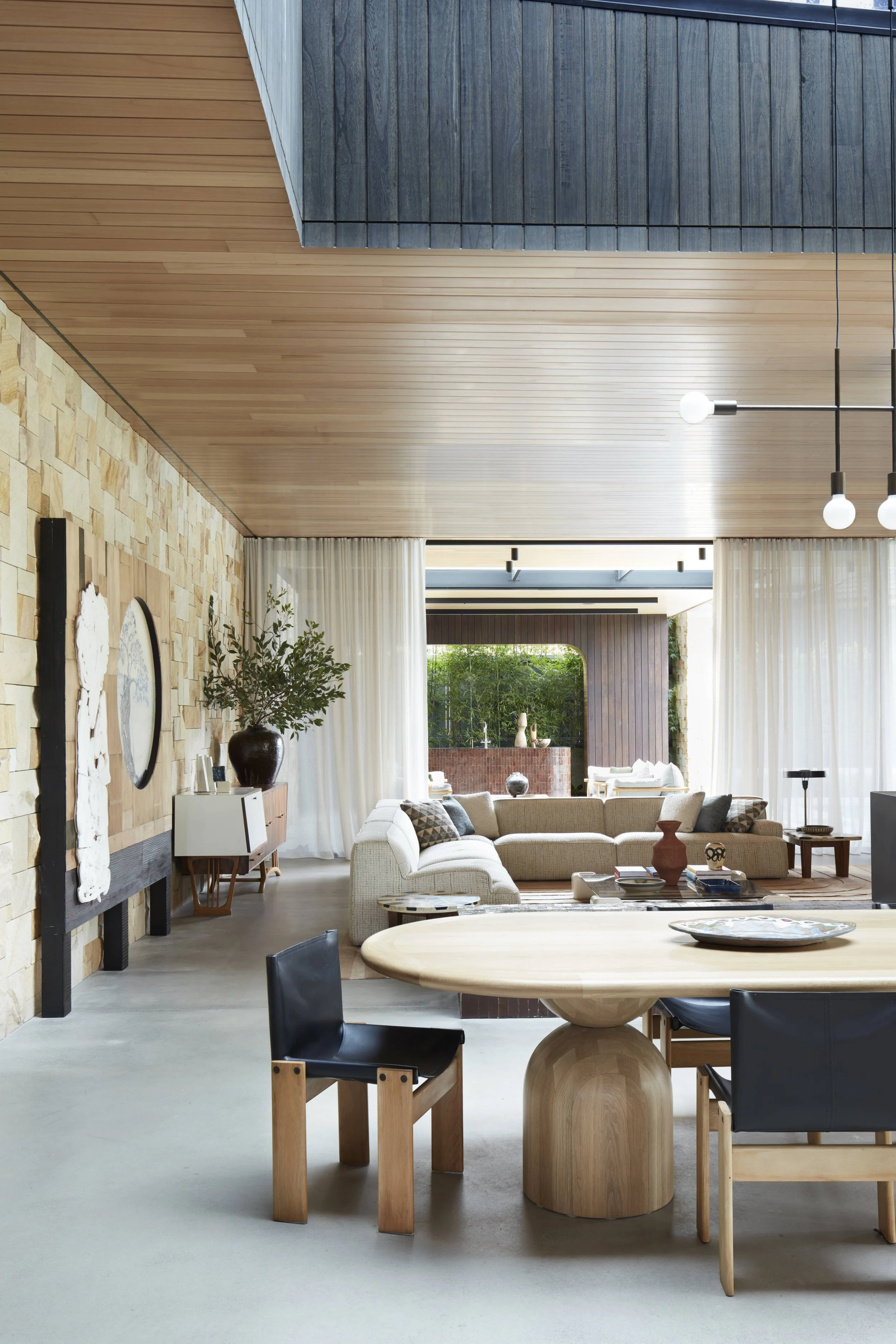 A spacious living room with a modern, rustic design. Features include a stone accent wall, a large beige sectional sofa with throw pillows, a wooden coffee table with decorative items, black and wooden chairs around a round wooden dining table, a con