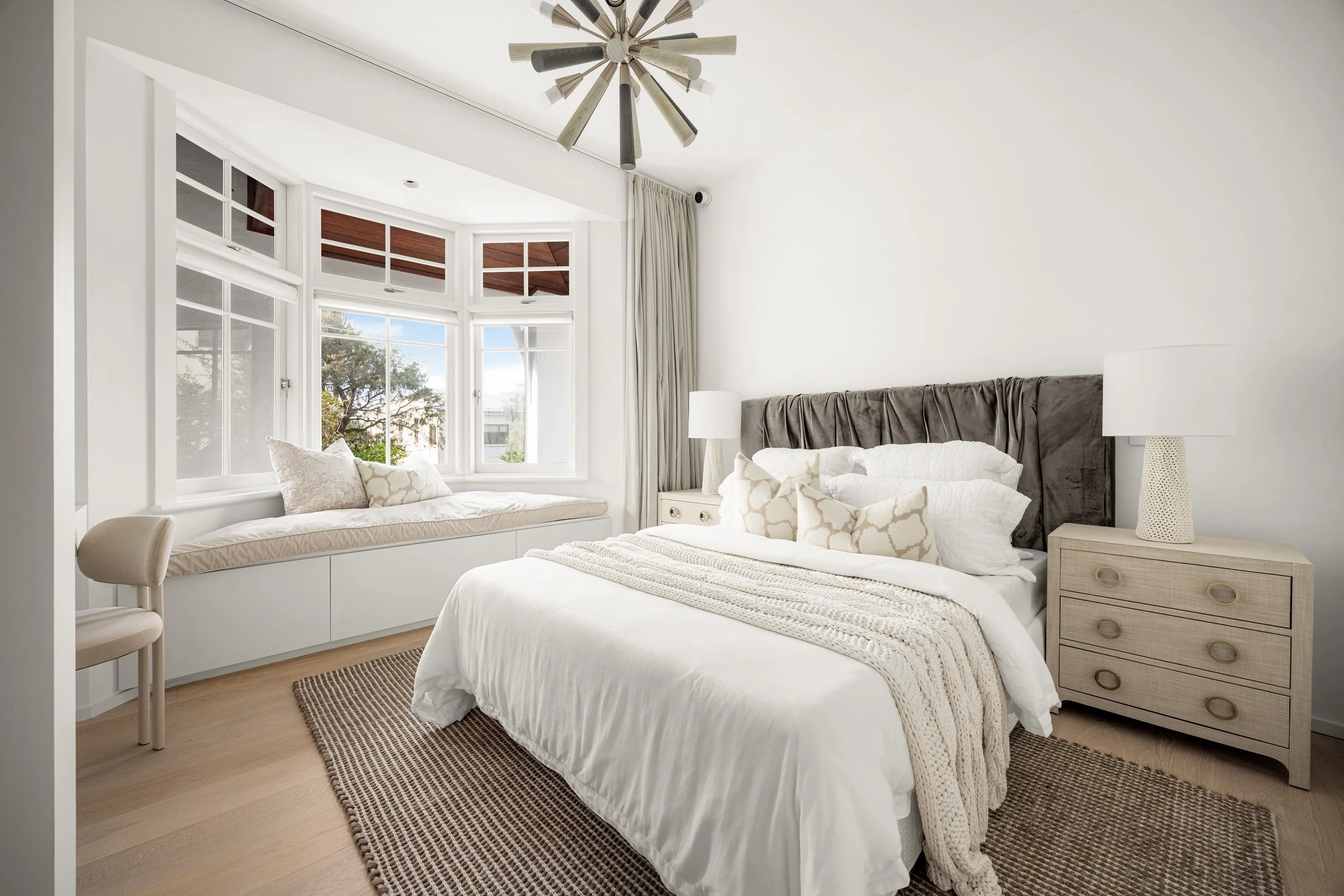 A bright bedroom with a large window seat, a bed with white linens and pillows, a bedside table with a lamp, and a modern chandelier on the ceiling.