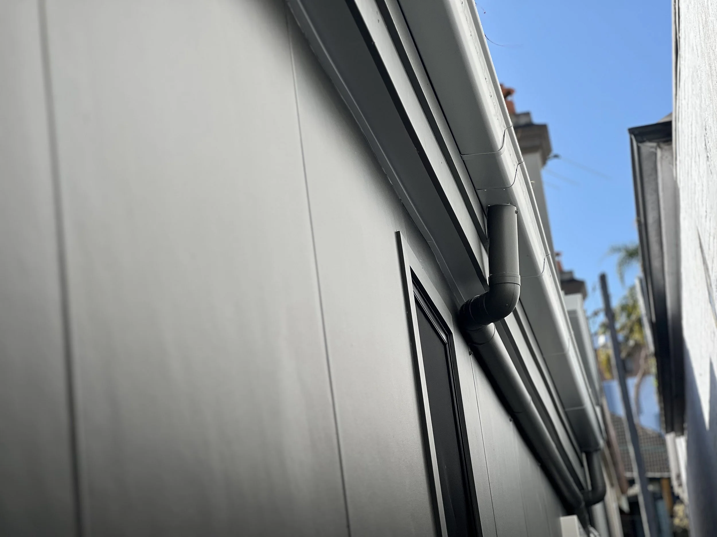 Close-up of a house gutter with black downspout, white fascia, and a partial view of neighboring houses against a clear blue sky.