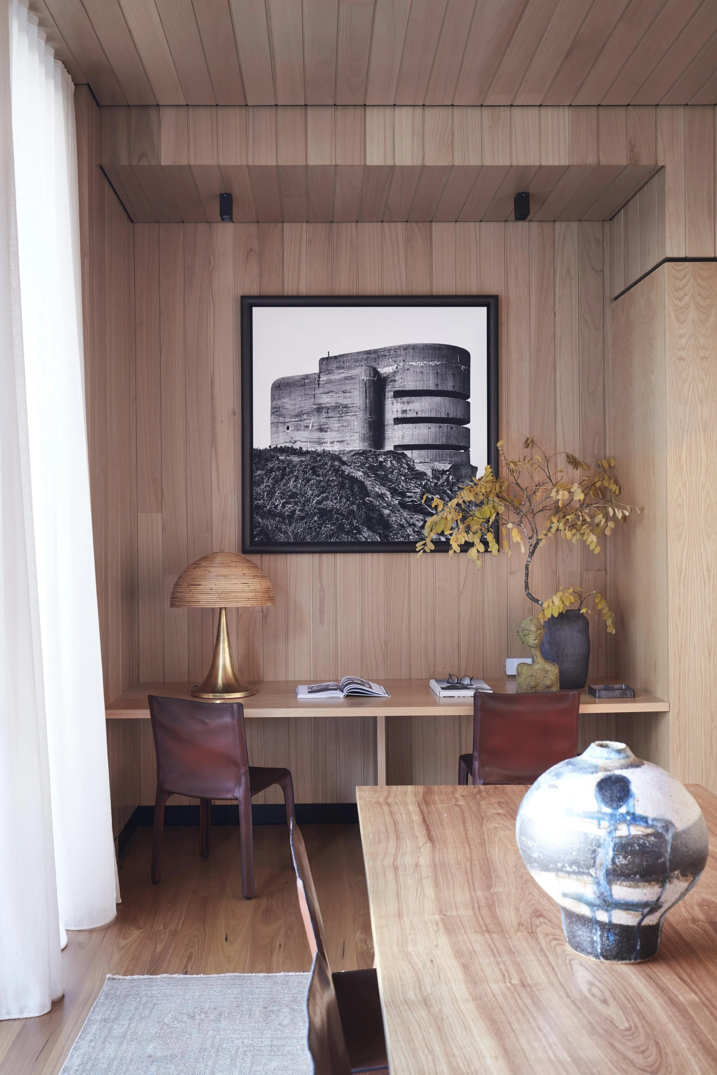 A cozy room with wood-paneled walls and ceiling, featuring a framed black-and-white photo of a modern concrete building, a desk with an open book, eyeglasses, a sculpture of a human head, and a vase with yellow leaves, complemented by a table lamp an